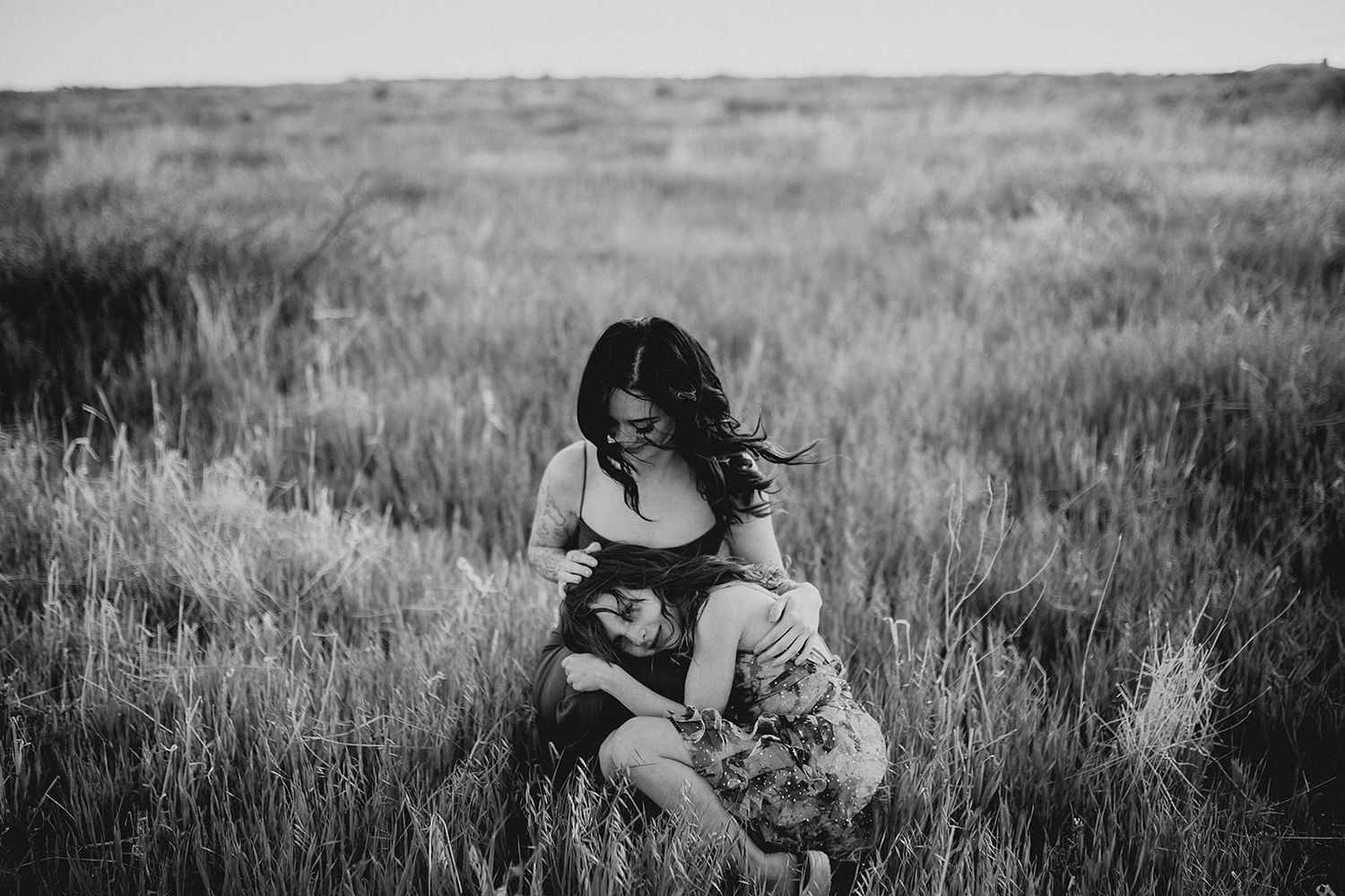 Black and white photo of a woman kneeling in a field, tenderly embracing a child among tall grass.