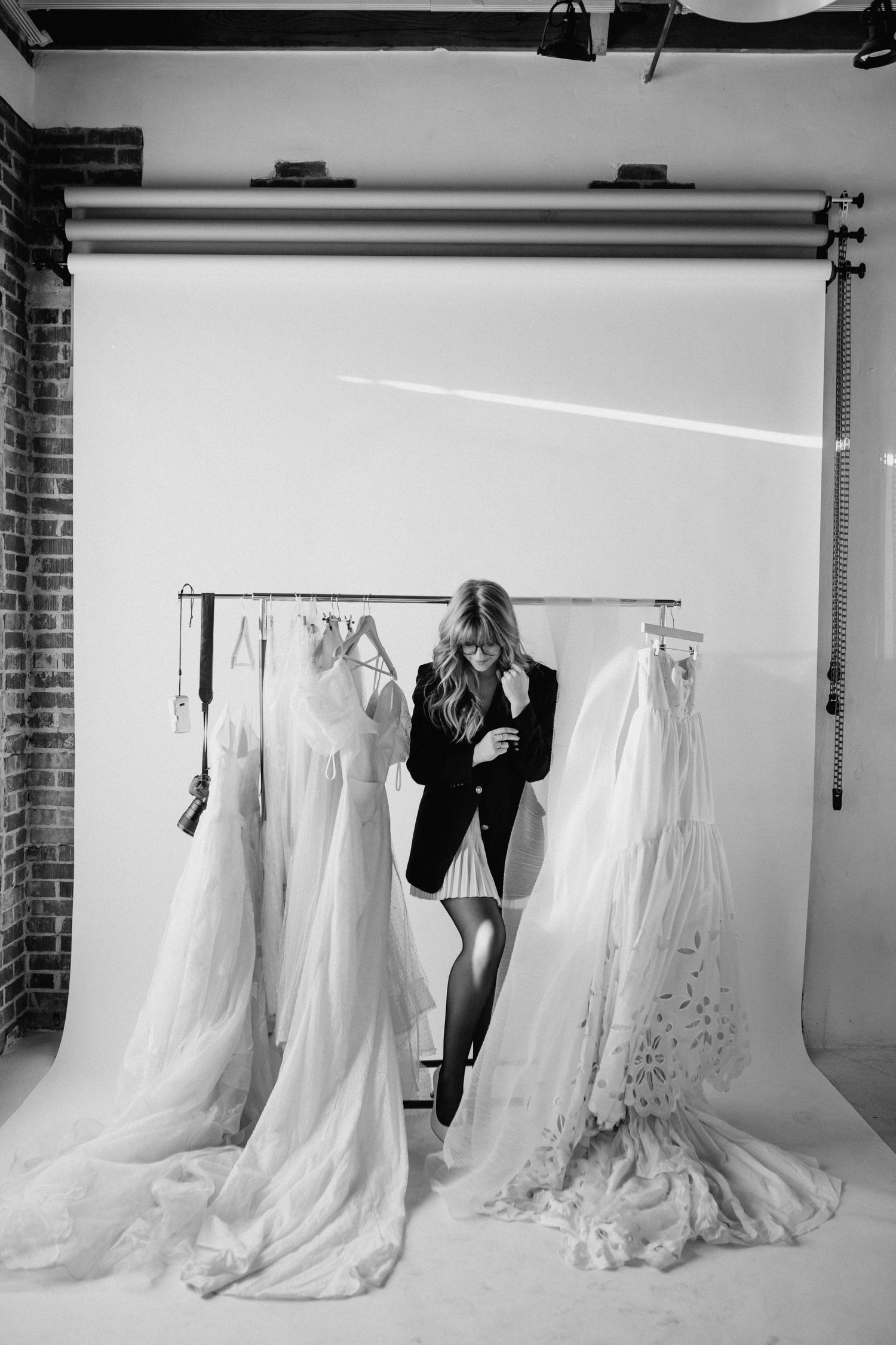 Woman browsing elegant white wedding gowns on a rack in a photography studio, black and white photo.