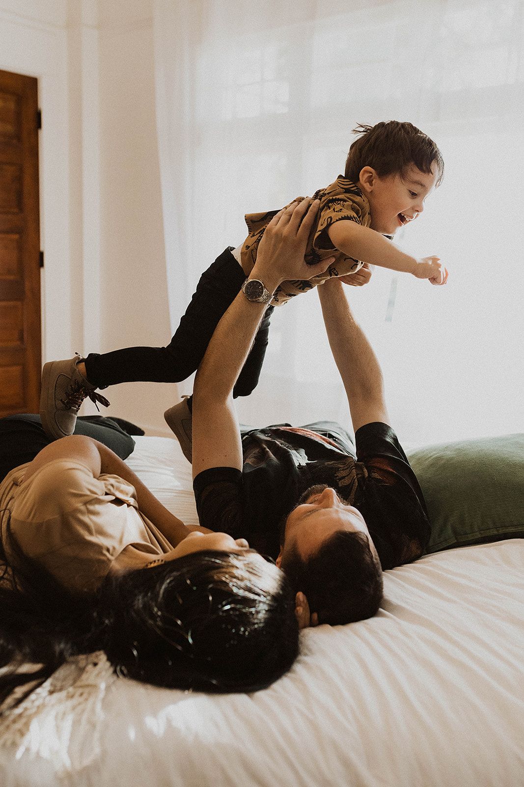 Parents lying on bed playfully lifting their young child in the air, enjoying a fun family moment indoors.