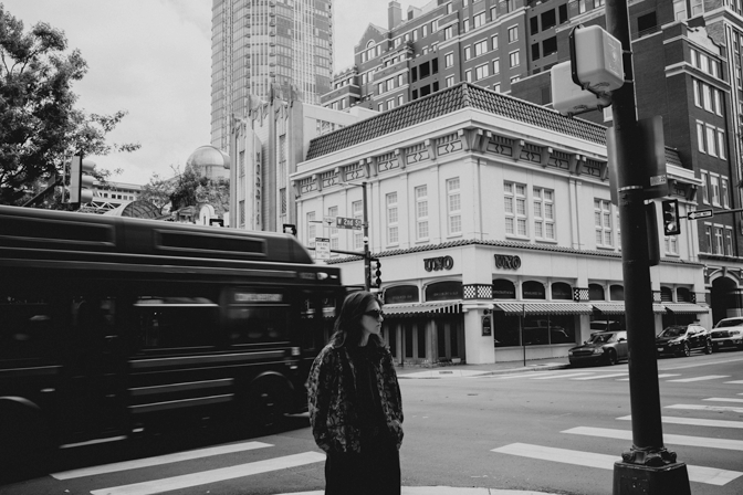 Black and white urban street scene with a person standing at a crosswalk, surrounded by classic city buildings and a passing bus.