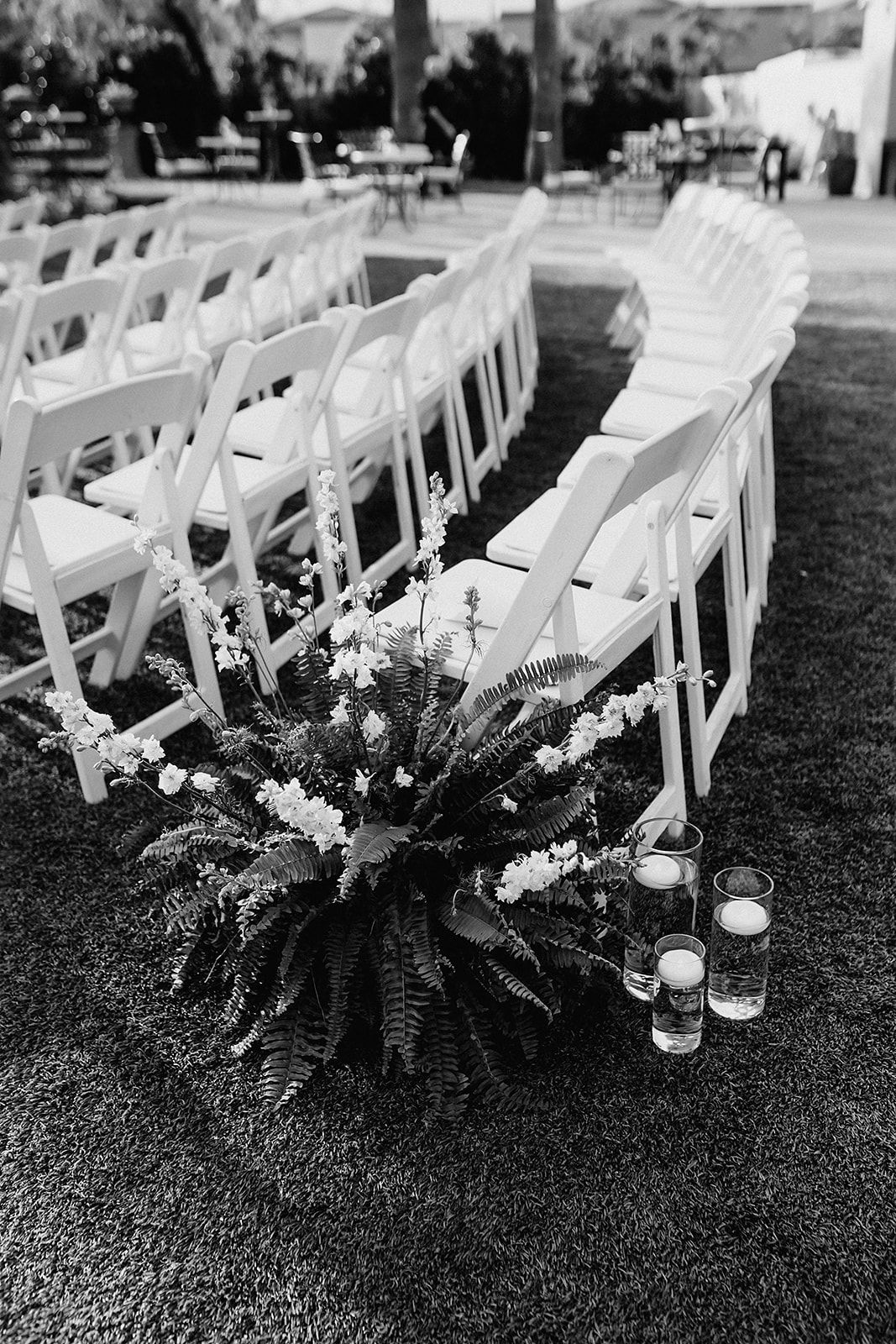 Black and white photo of white wedding ceremony chairs arranged in rows with floral arrangement and candles on grass.