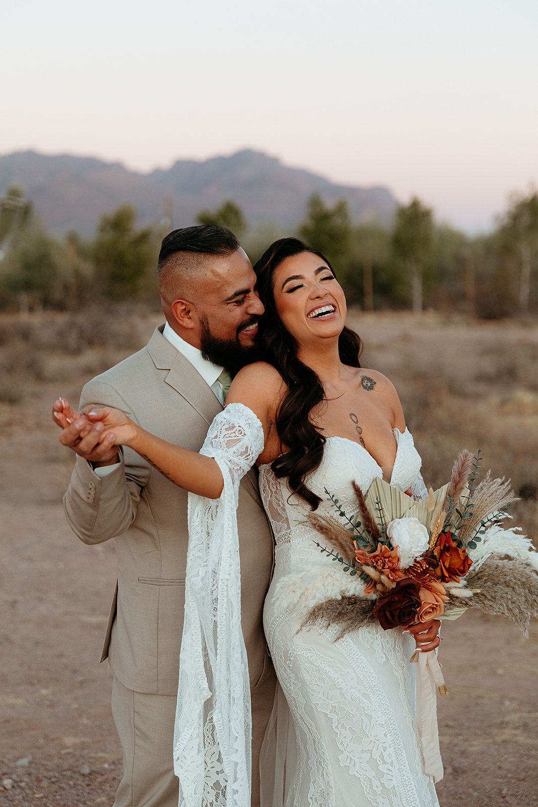 A joyful couple shares a candid moment at their desert wedding with mountains in the background at sunset.