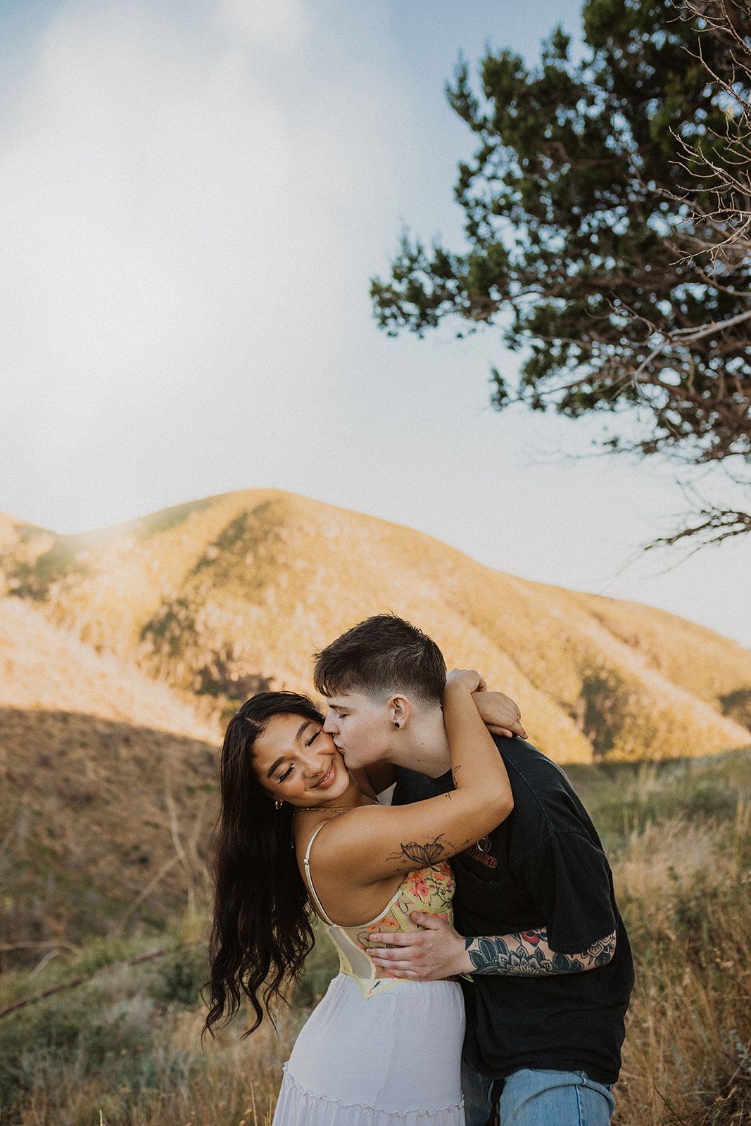 A couple shares a romantic embrace against a backdrop of golden hills and mountains during sunset.