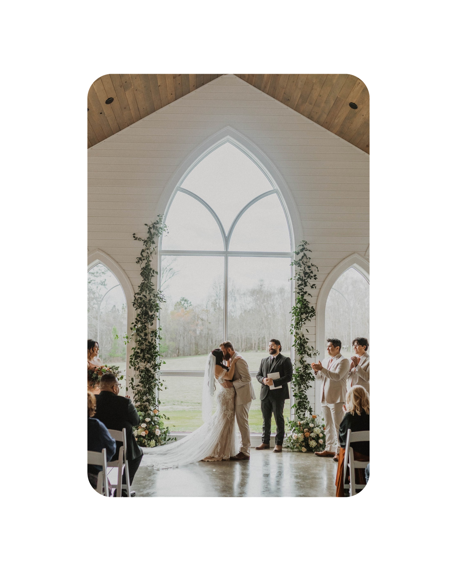 A wedding ceremony takes place in a bright chapel with gothic arched windows and greenery-adorned columns.