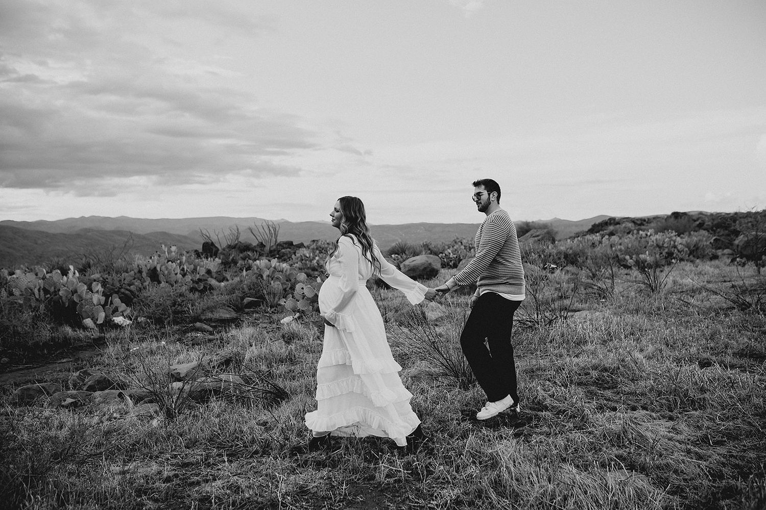 A couple in wedding attire walks through a scenic grassy hillside landscape in black and white.