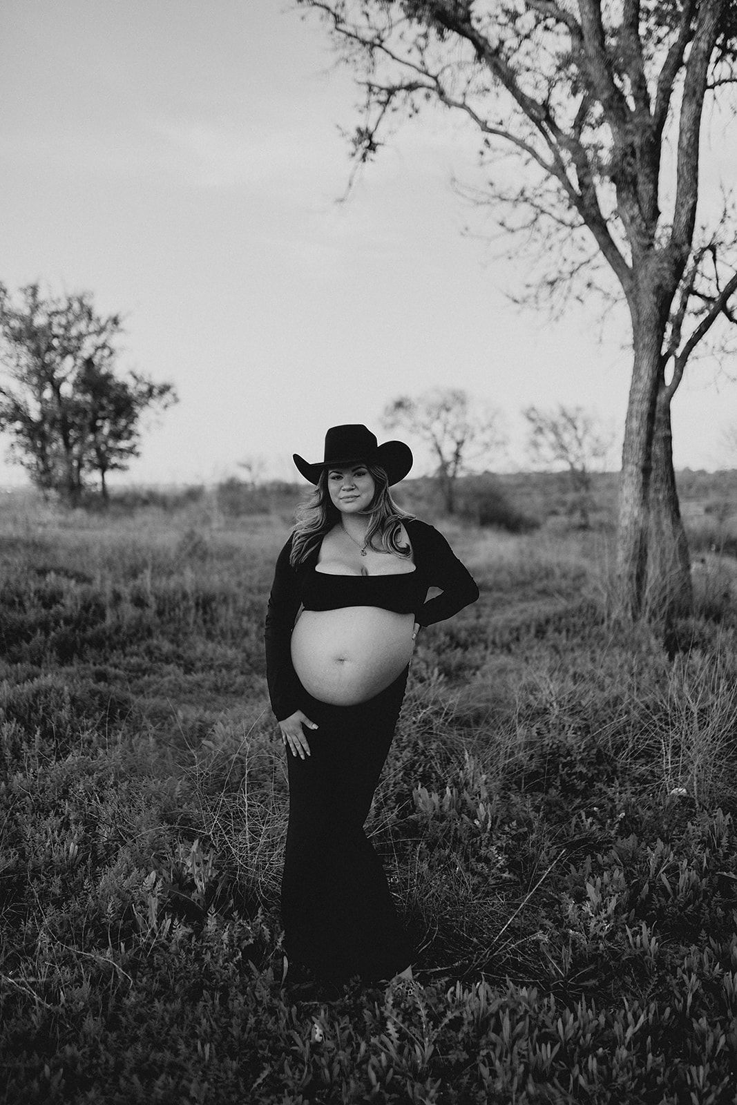 A black and white maternity photo shows a person in a cowboy hat and long dress posing outdoors in a rustic field setting.