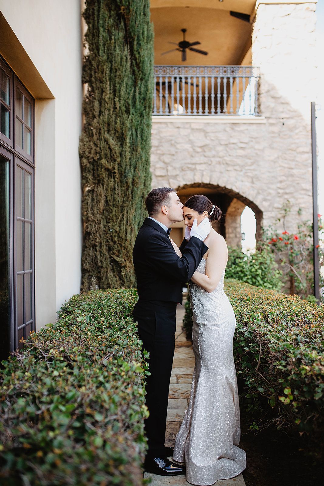 Couple shares intimate moments during wedding photoshoot in front of stone wall and cypress trees.