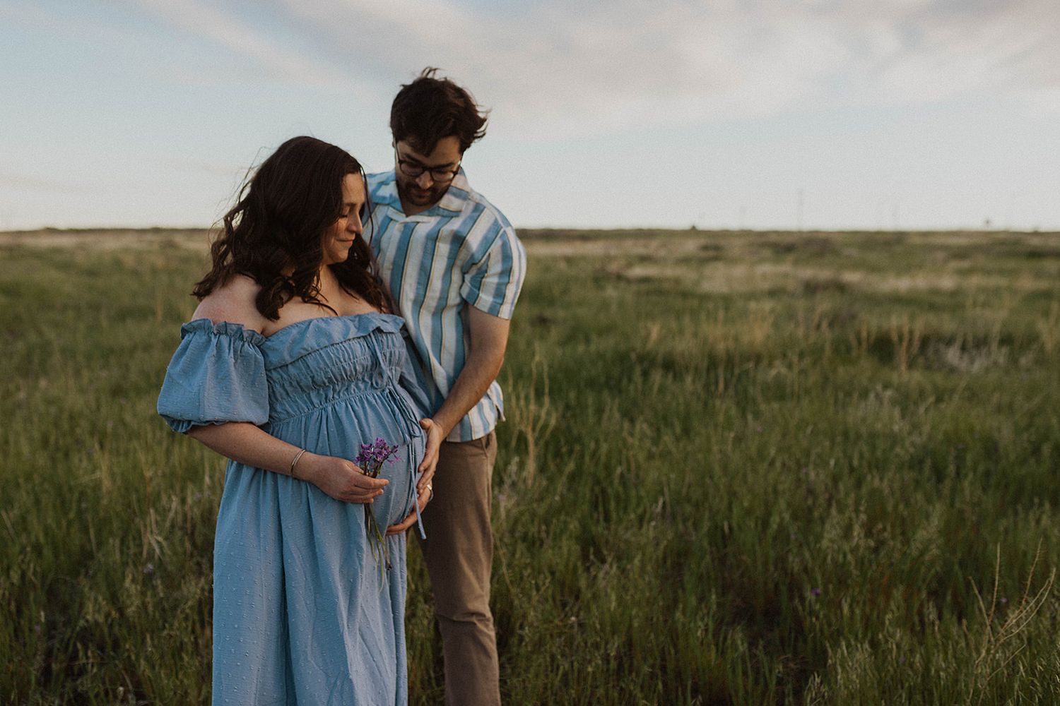 A couple in casual attire embraces while standing in a vast grassy field during golden hour.