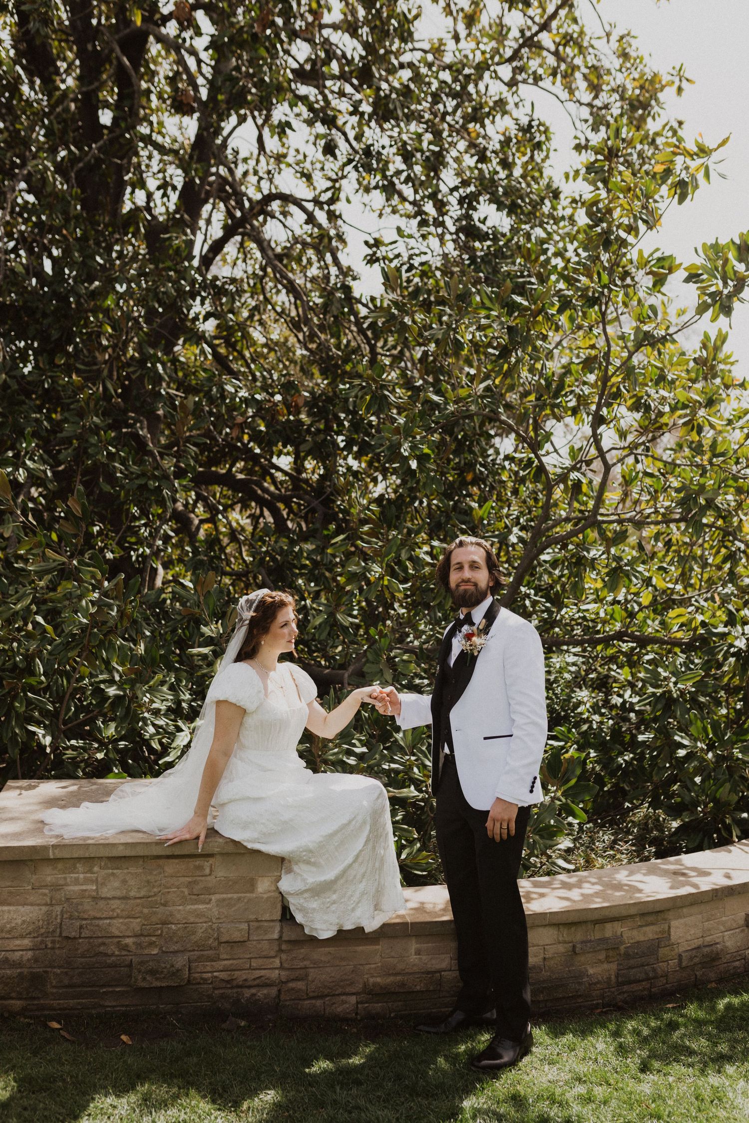 Couple in wedding attire poses playfully on a rustic wooden wall surrounded by lush greenery on a sunny day.