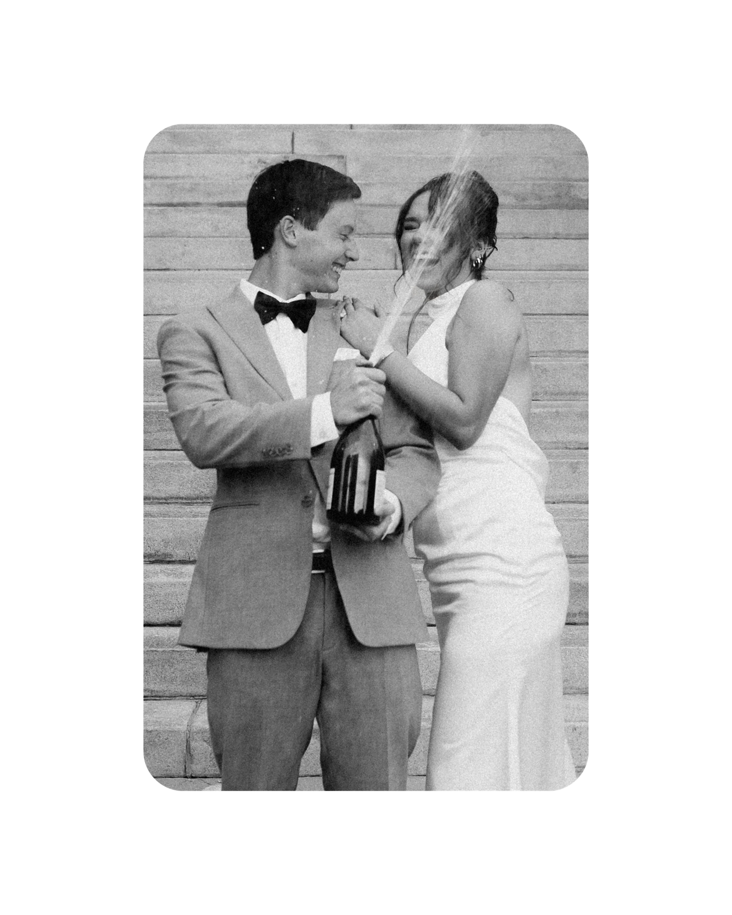 A black and white wedding photograph showing a couple in formal attire sharing a tender moment against a wall.