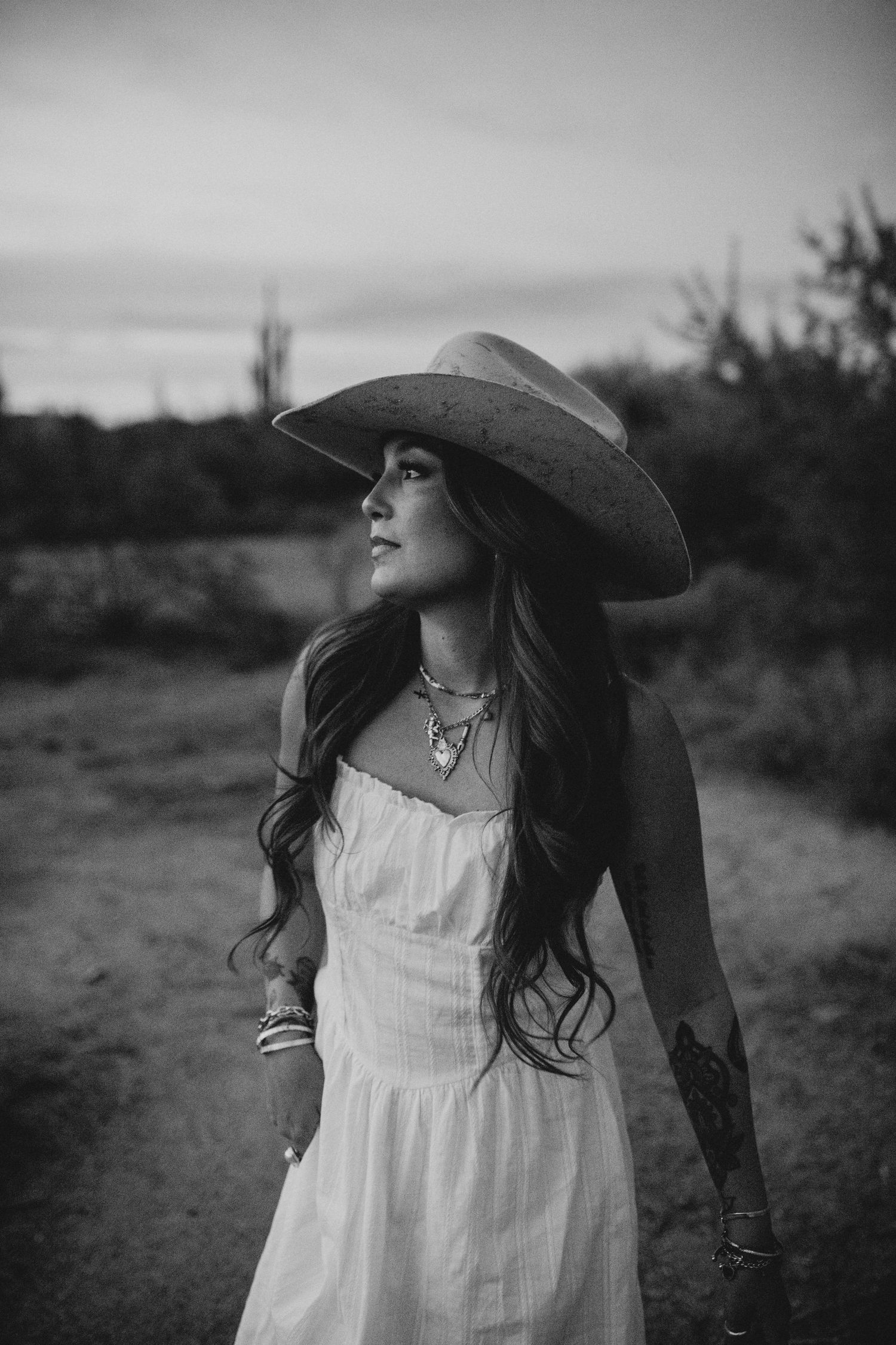 Black and white photo of a woman in a white dress and wide-brim cowboy hat standing outdoors at dusk.