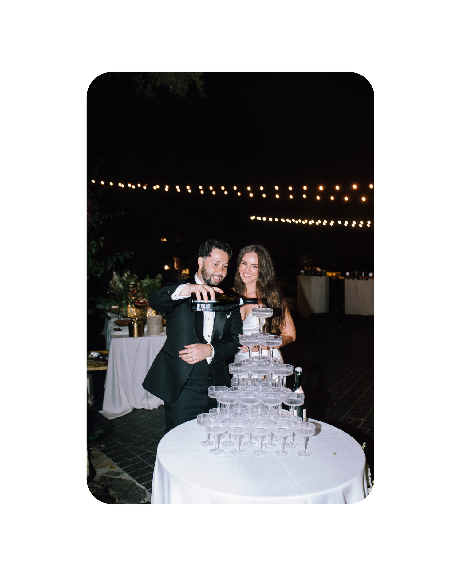 A couple stands behind a tiered champagne tower at an outdoor evening reception with string lights overhead.