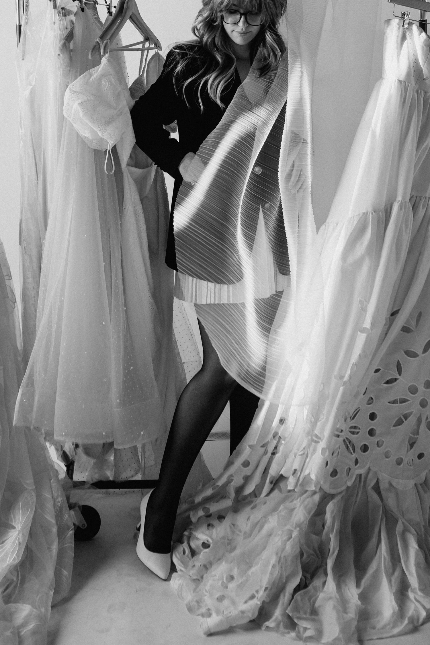 Black and white photo of a woman in a structured dress surrounded by elegant white gowns hanging on racks in a bridal boutique.