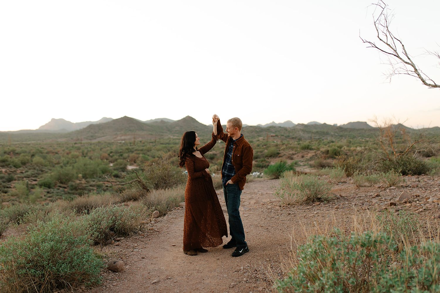 Two people in casual attire dance together on a desert trail surrounded by sparse vegetation and rolling hills.