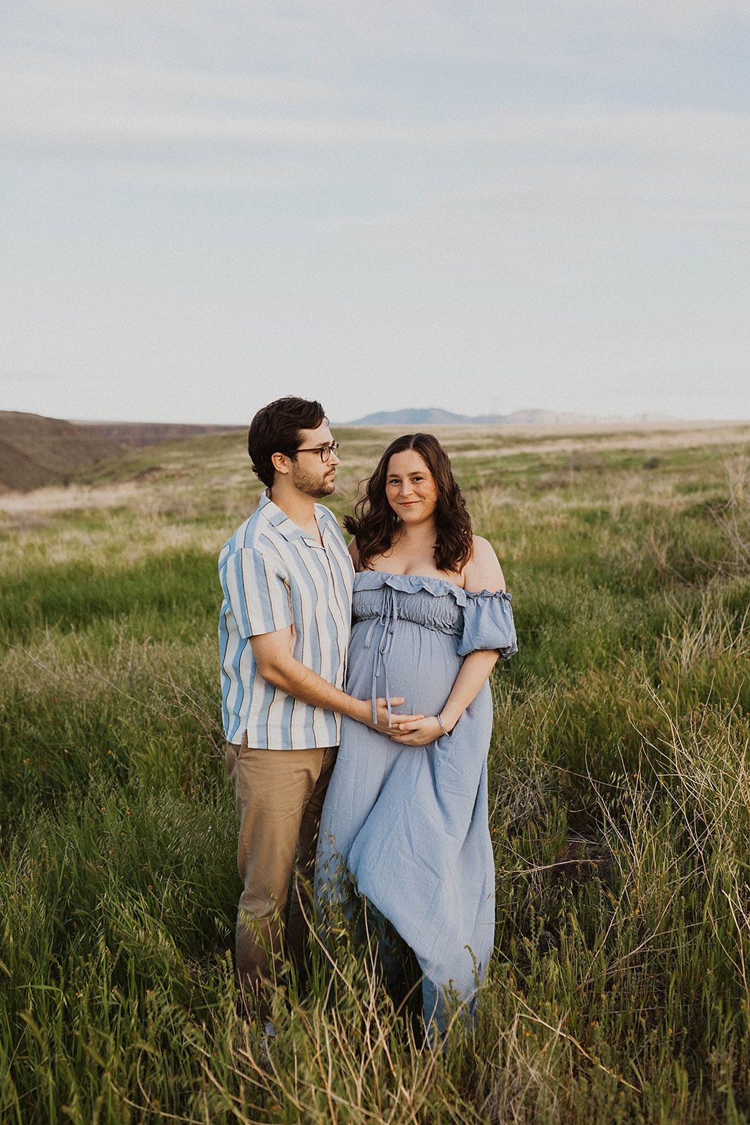 A couple stands together in a grassy field during golden hour, with mountains visible in the background.