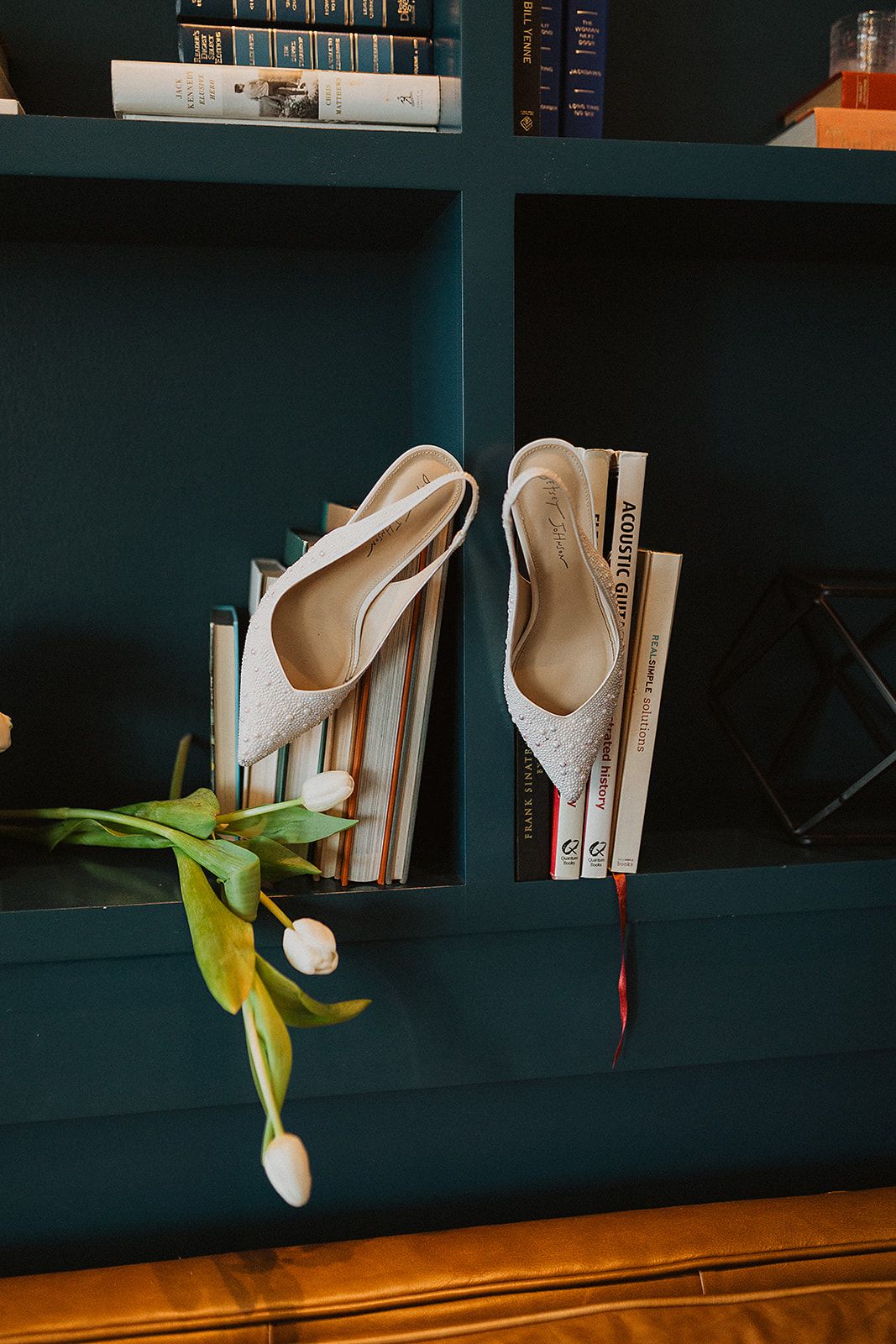 White bridal shoes rest on books in a dark teal bookshelf with yellow tulips beside them.