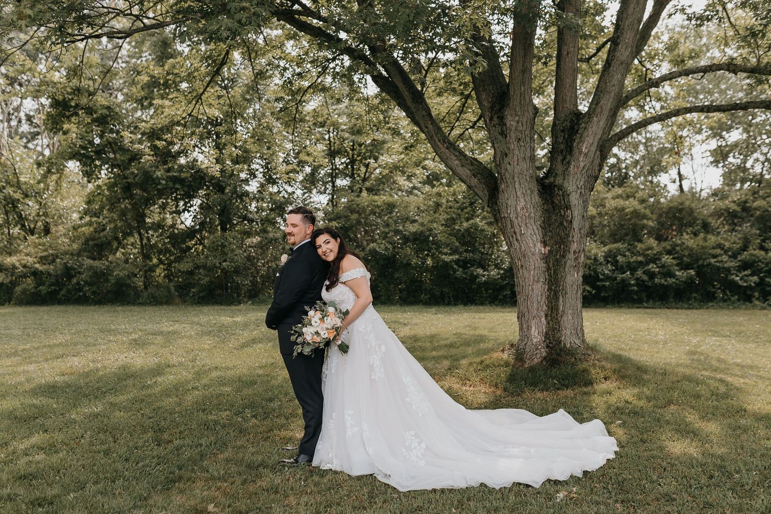 A newlywed couple poses together under a large tree on their wedding day.
