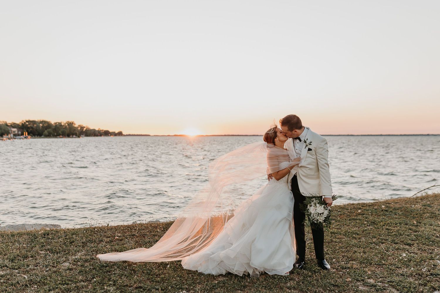 Couple embraces at sunset by the water with the bride's dress flowing in the breeze.