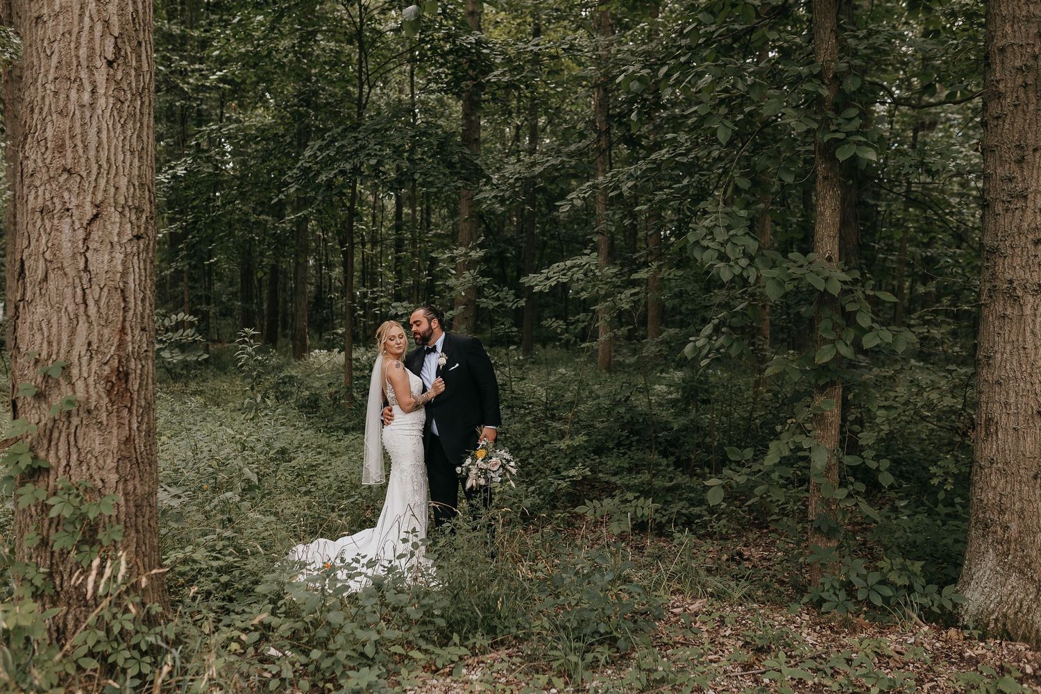 A couple in wedding attire embrace in a serene forest setting with tall trees and natural greenery.