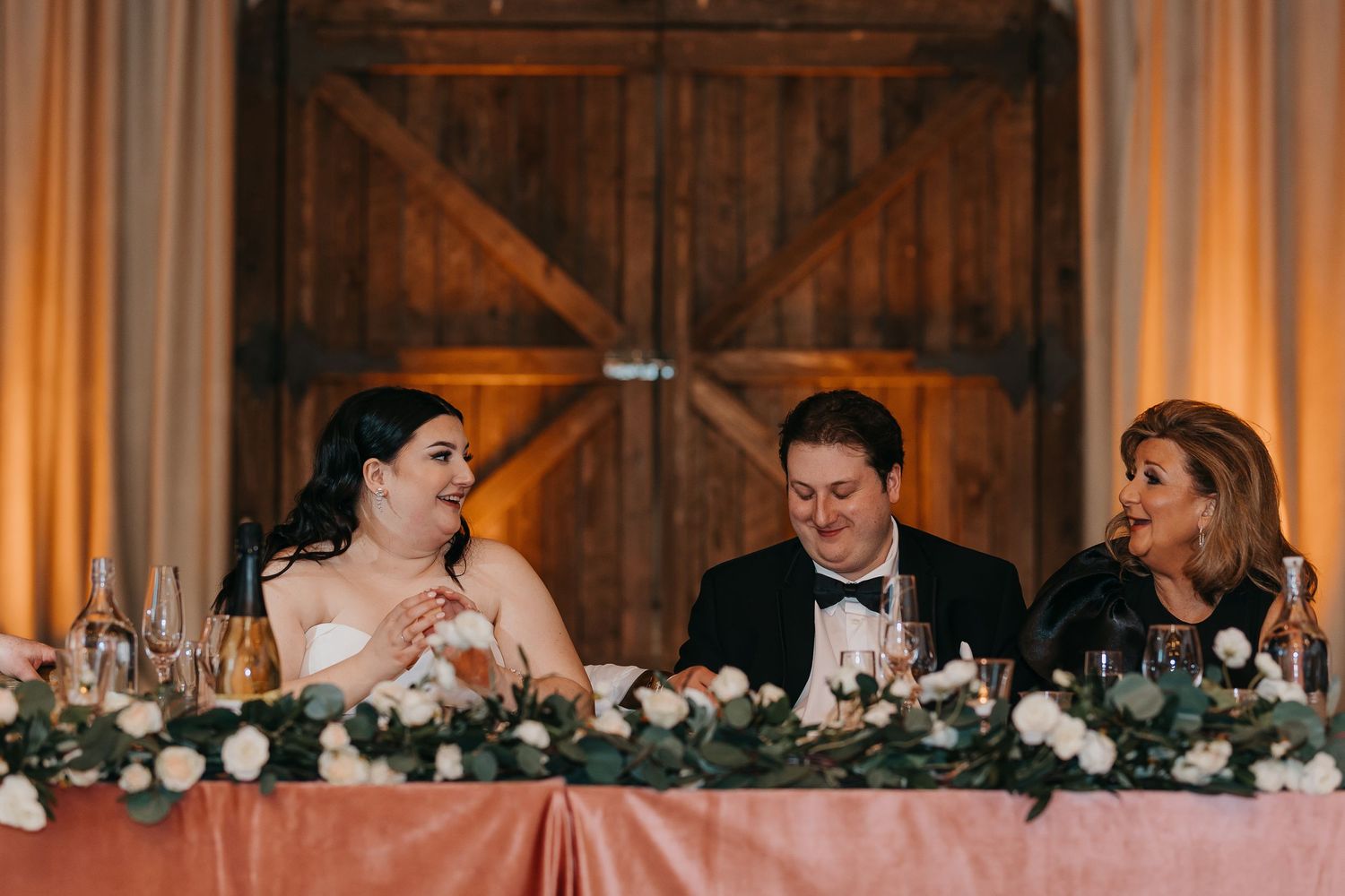 Wedding reception scene with guests seated at a head table decorated with greenery and candles.