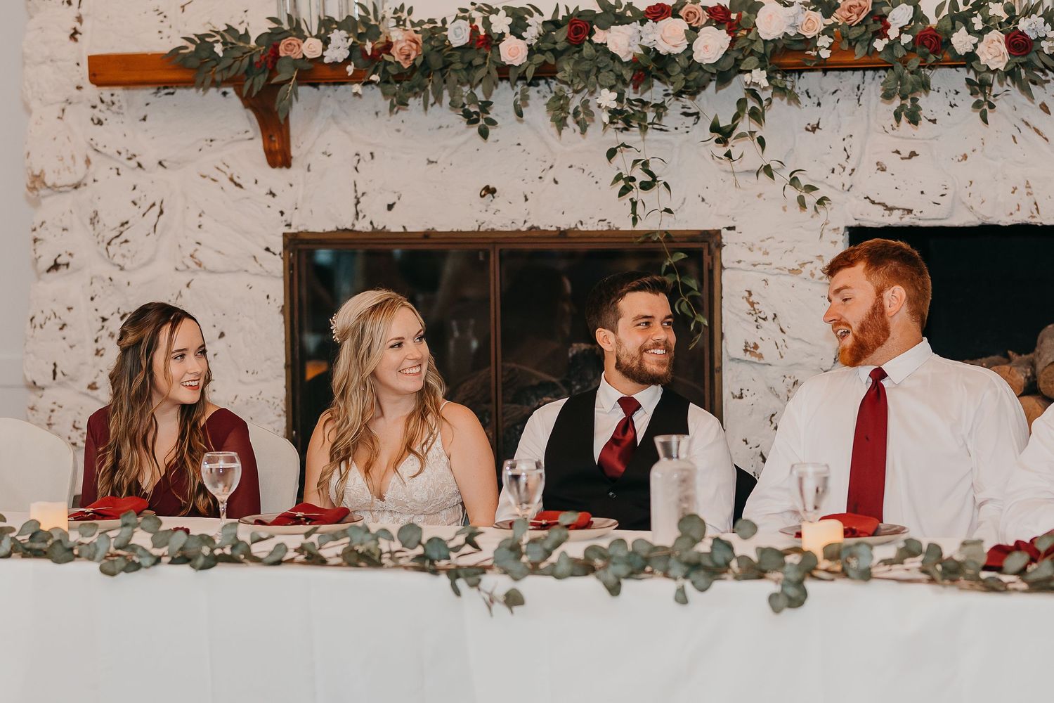 Wedding party seated at a rustic head table decorated with greenery and flowers during reception.