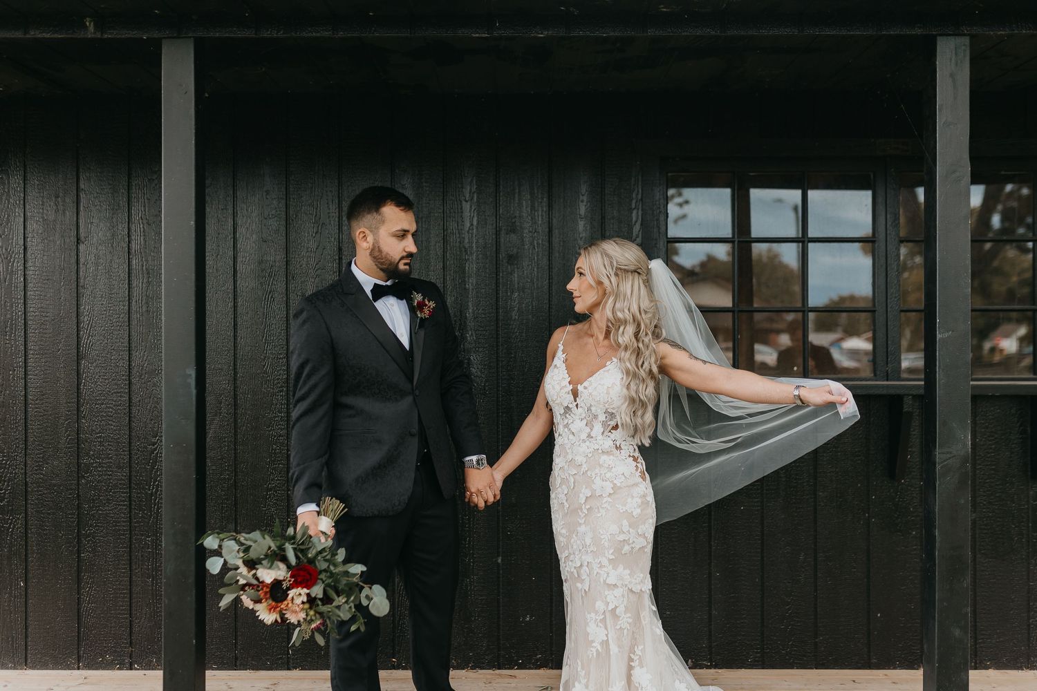 A wedding couple poses against a dark wooden building exterior with floral arrangements.