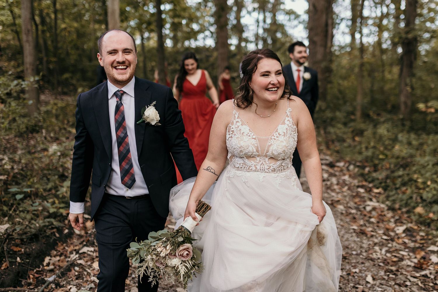 Newlyweds walk through fallen leaves on woodland path during autumn wedding.