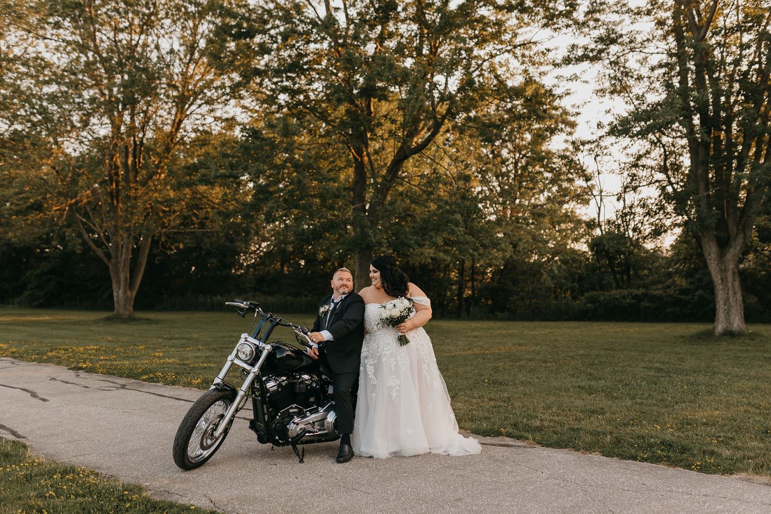Couple poses with a motorcycle on their wedding day in a scenic outdoor setting.