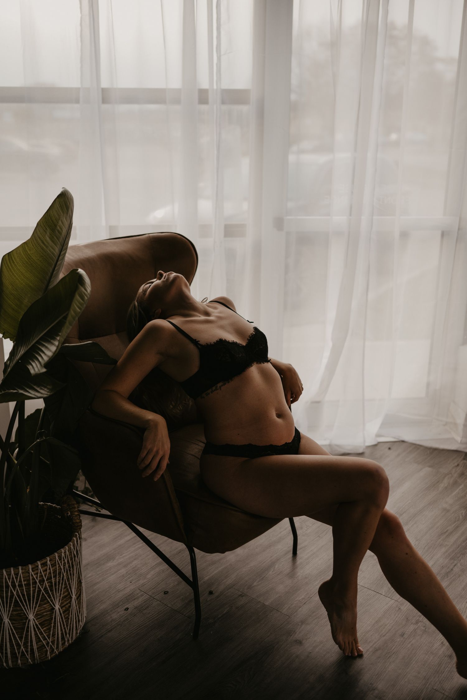 Woman in black lingerie reclining in a leather chair by sheer curtains with a tropical plant nearby.