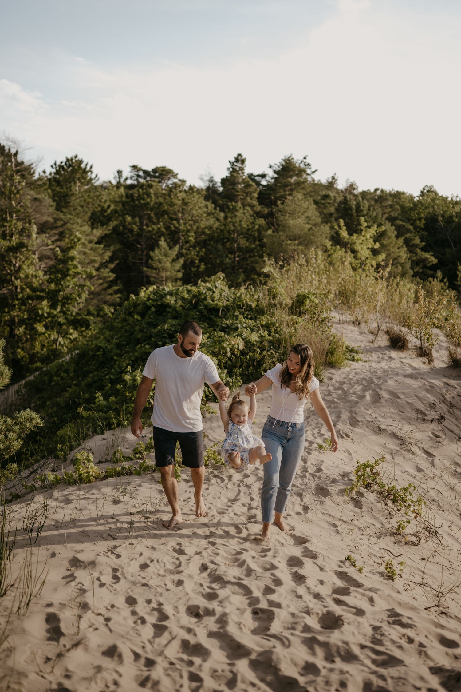 Parents swinging toddler between them while walking on sandy beach path with lush green trees behind.