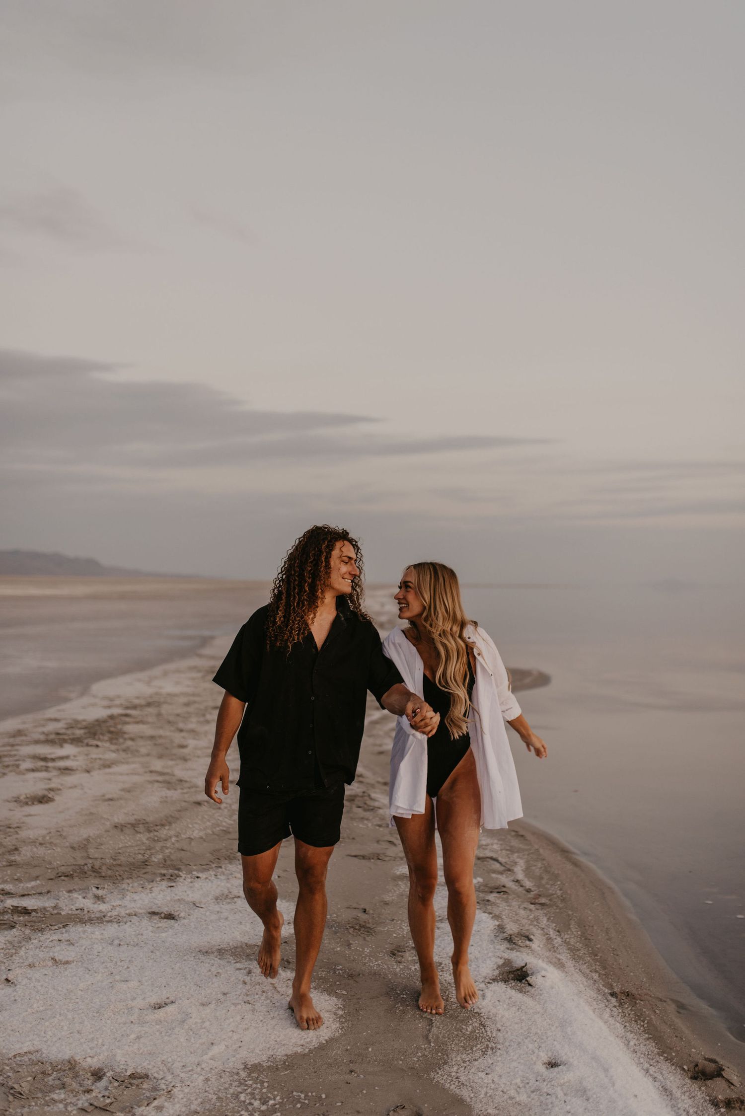 Couple walking barefoot along the Great Salt Lake shoreline at dusk holding hands.