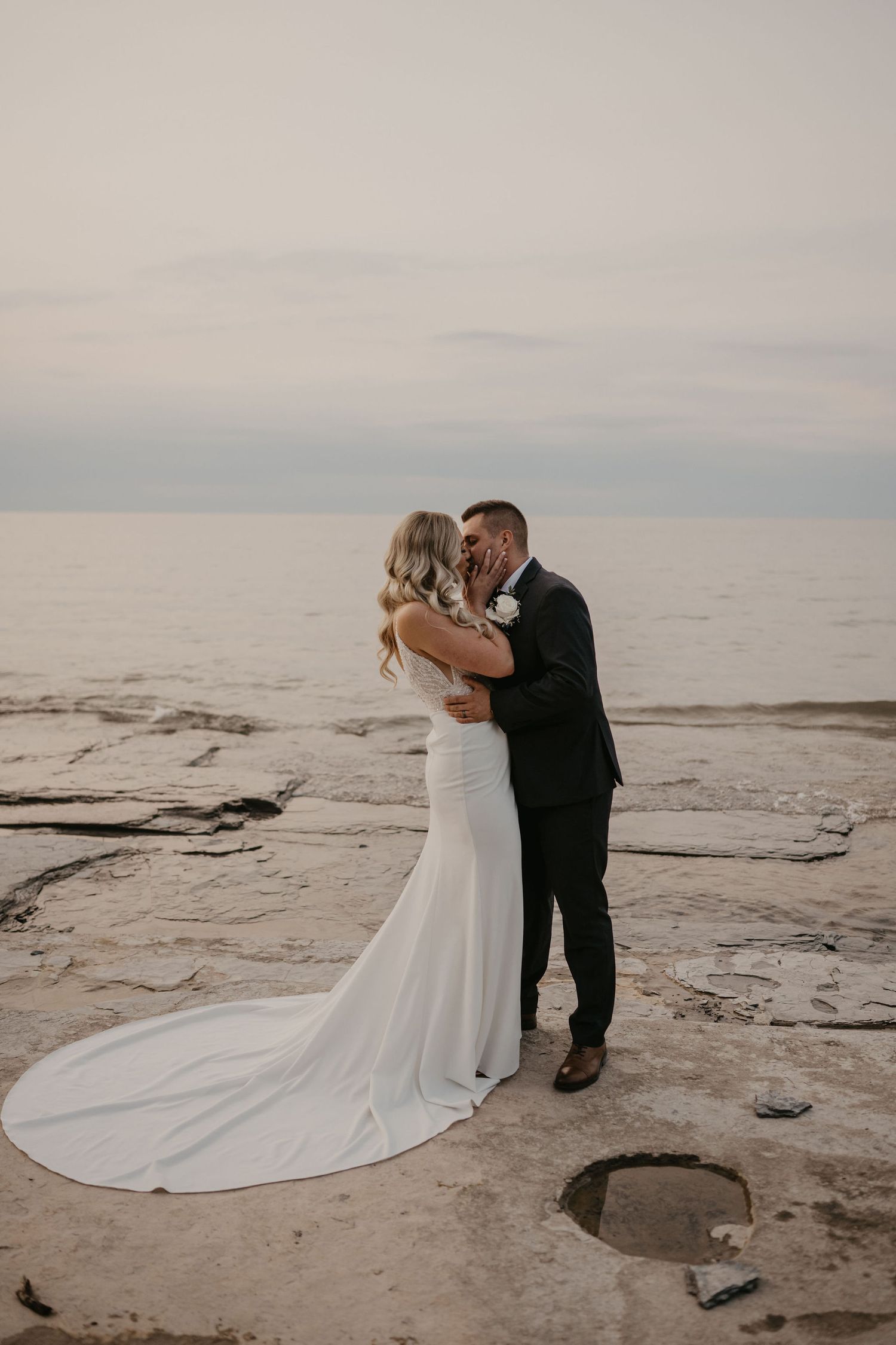 Bride and groom embrace romantically on rocky shoreline at dusk in elegant wedding attire.
