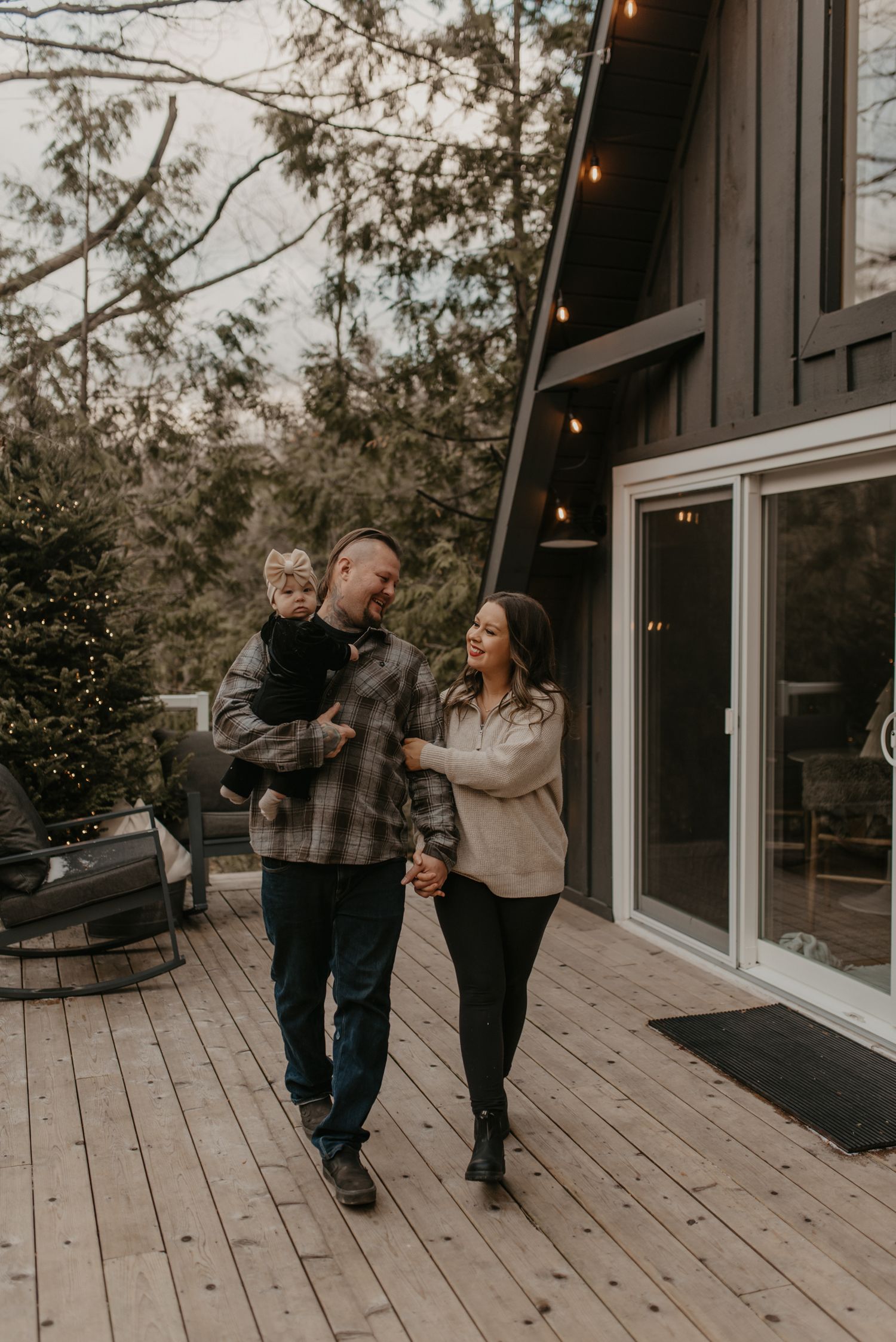 A happy family walks on a wooden deck outside a modern dark home, with a man holding a toddler and a woman beside him.