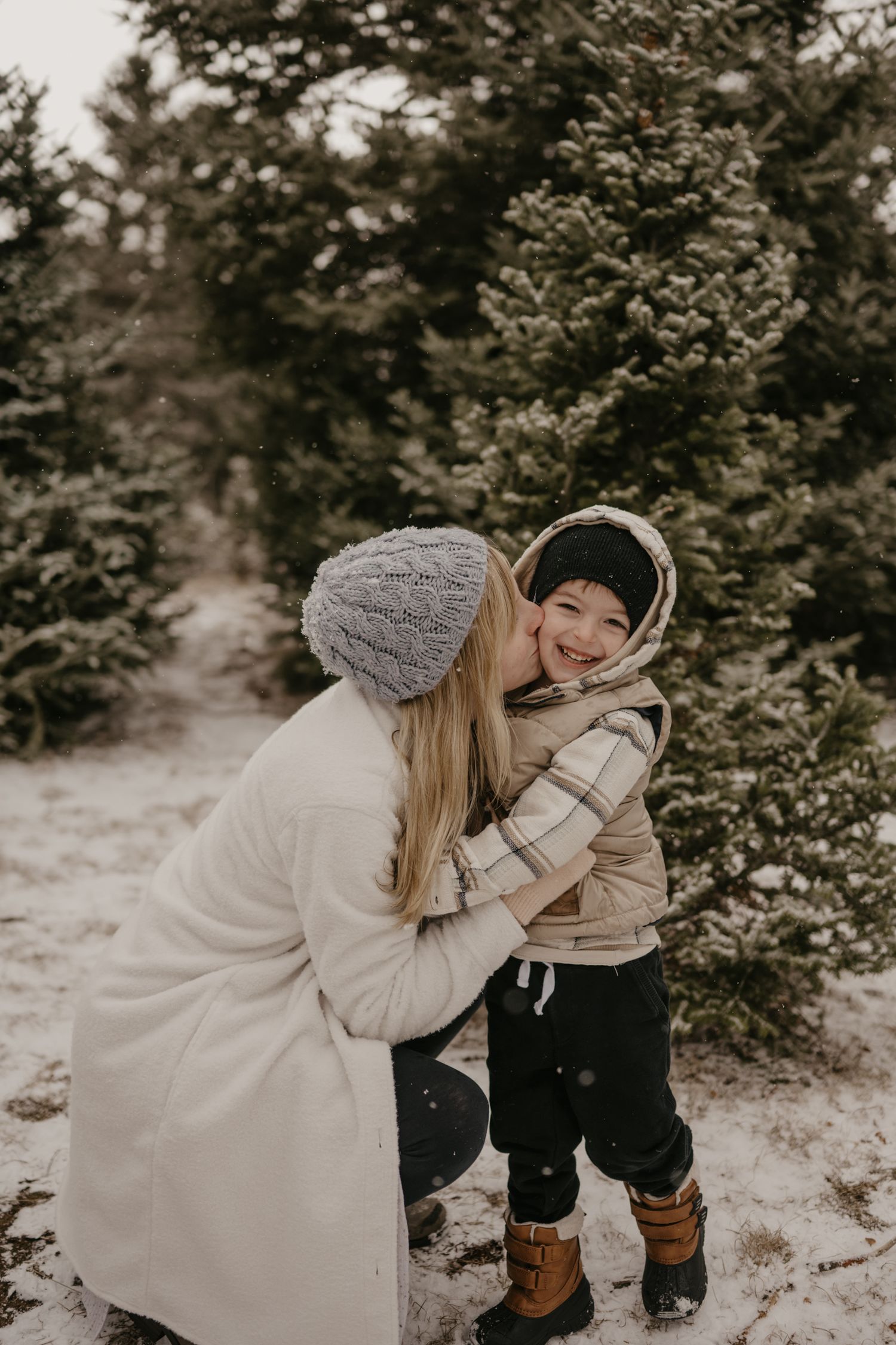 Mother kissing smiling child on cheek at snowy Christmas tree farm, both wearing winter coats and hats.