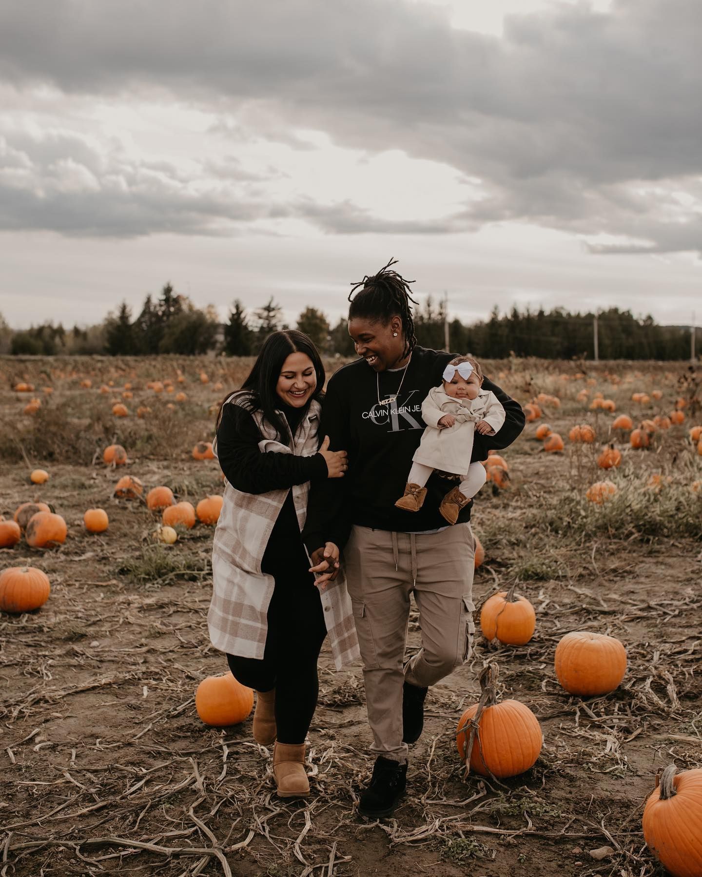 A couple smiles while holding their baby among orange pumpkins in a fall pumpkin patch under cloudy skies.