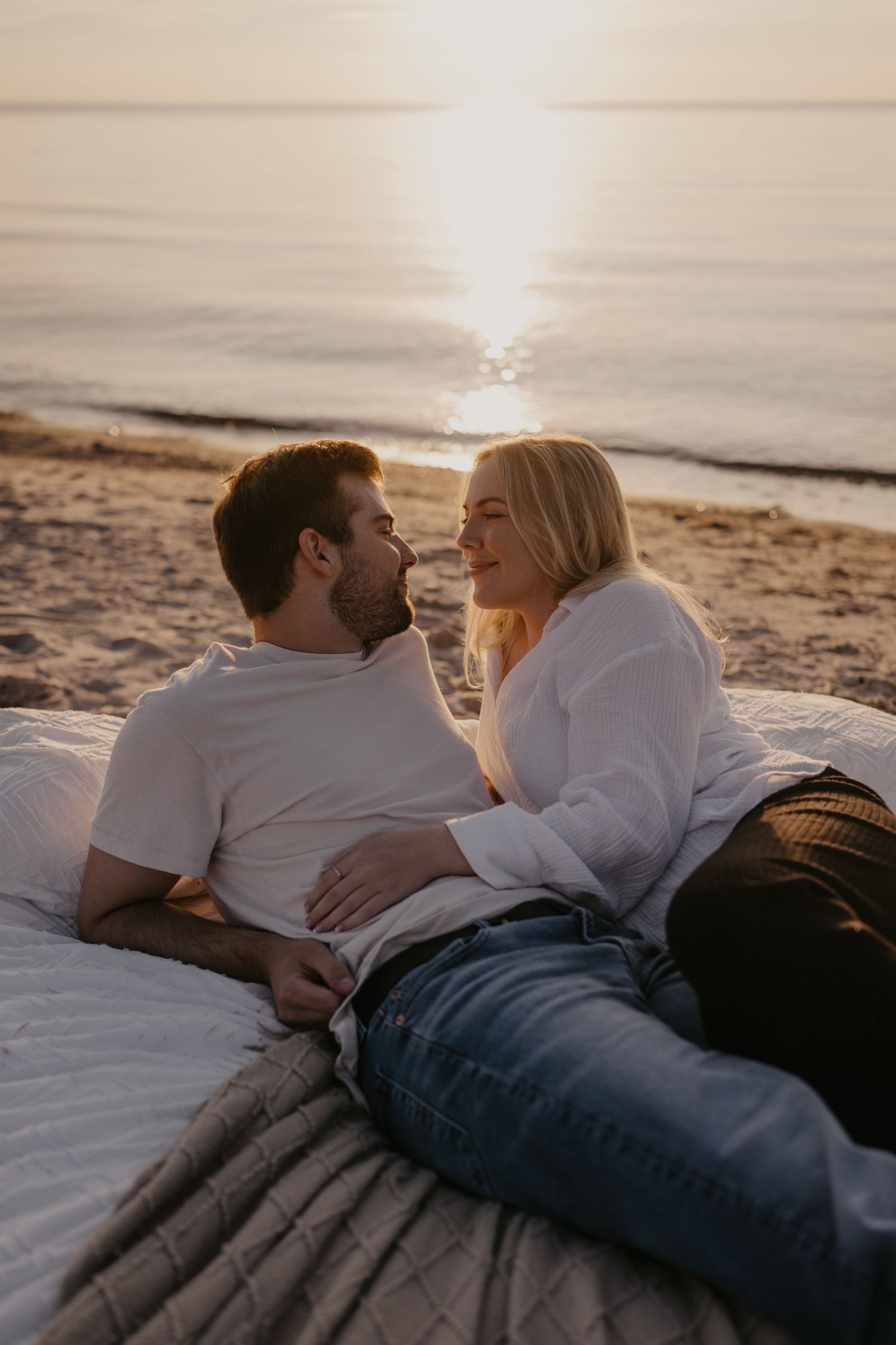 Happy couple lying on beach blanket at sunset, smiling and gazing into each other's eyes.