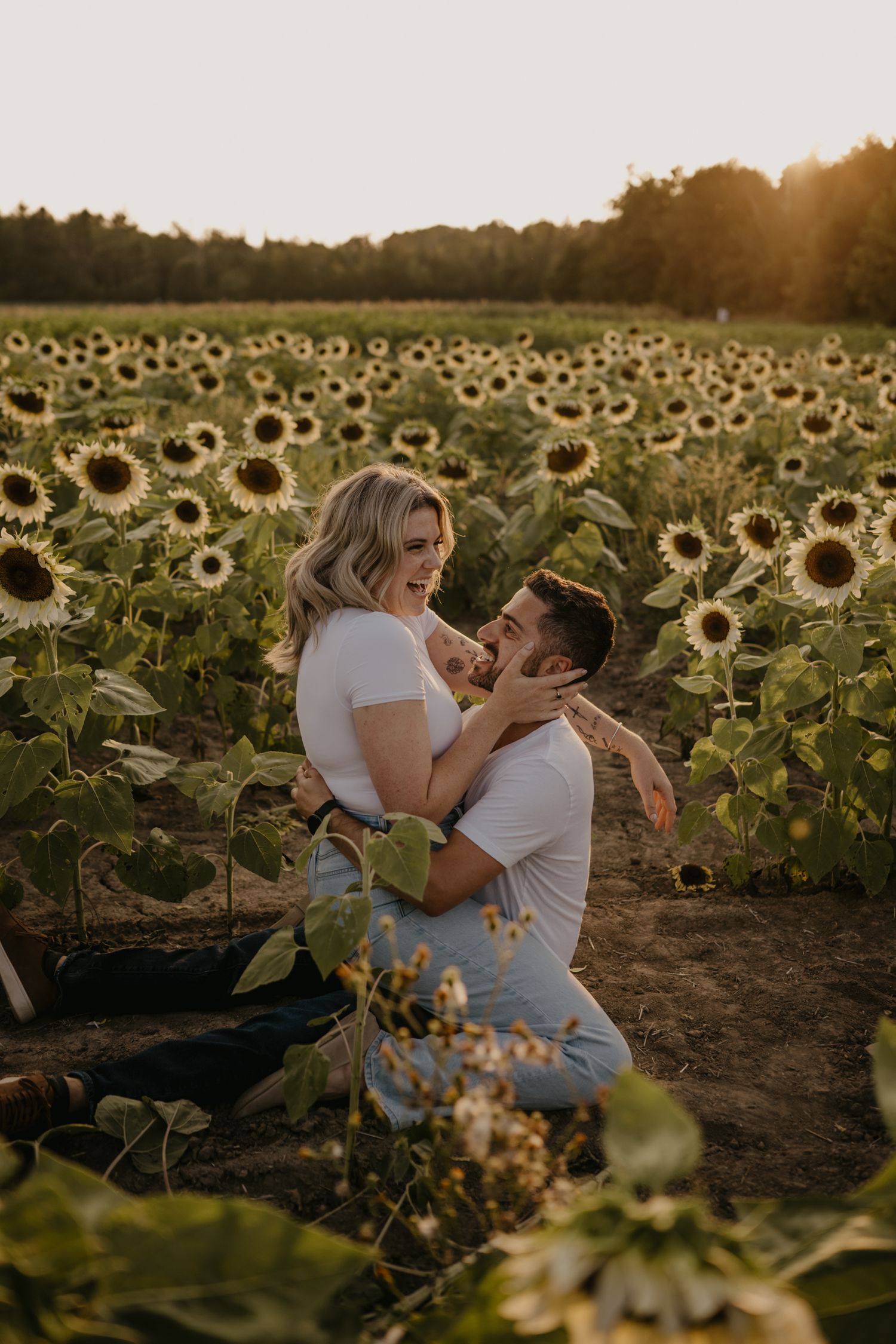 Man on one knee lifting and embracing laughing woman in sunflower field during warm sunset light.