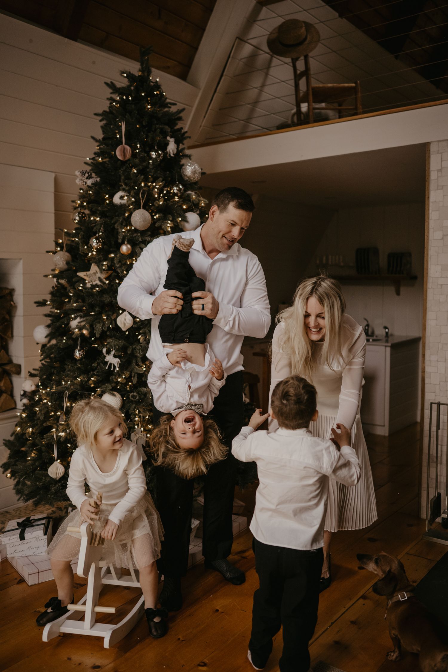 Family laughing together by decorated Christmas tree, dad holding child upside down, kids in white outfits, festive holiday photo.