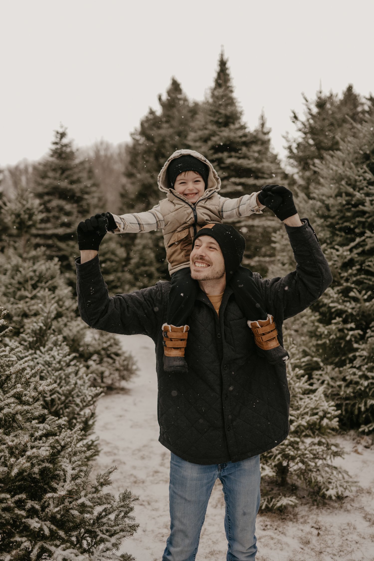 Father carries smiling child on shoulders at snowy Christmas tree farm in winter, both wearing warm coats and hats.