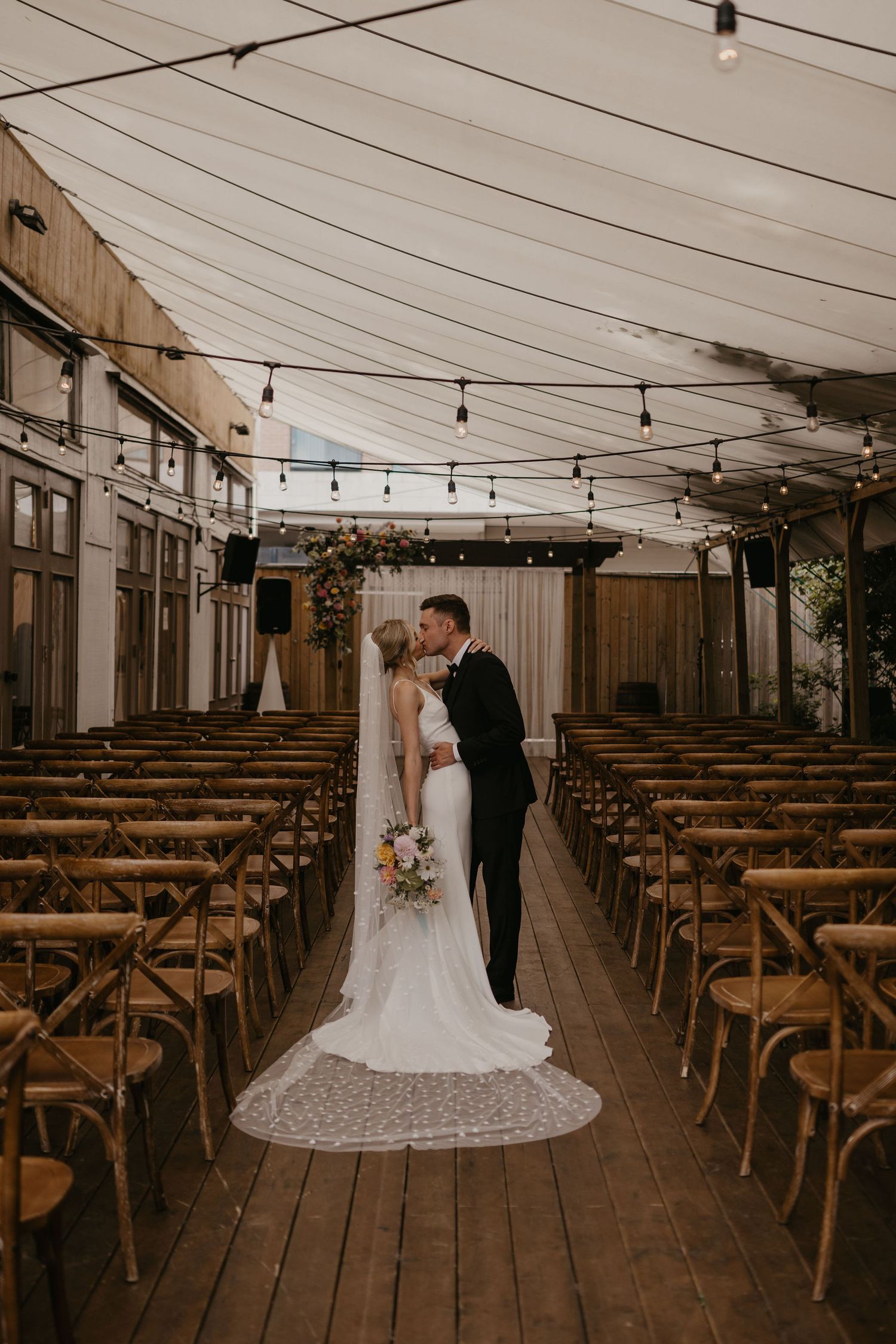 Newlyweds share first kiss at altar in rustic tent venue with string lights.