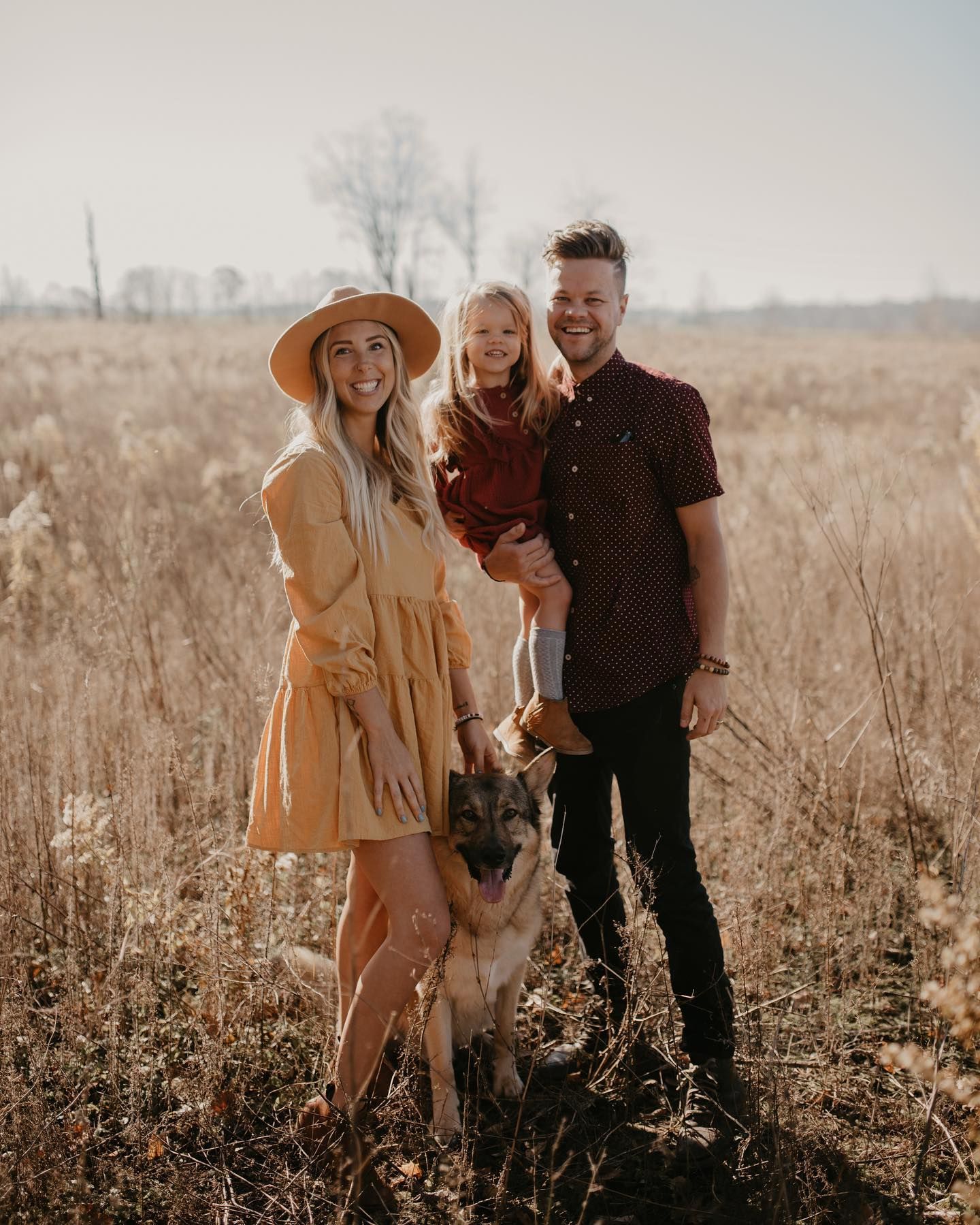 Family posing with German Shepherd dog in golden autumn field, woman in yellow dress.