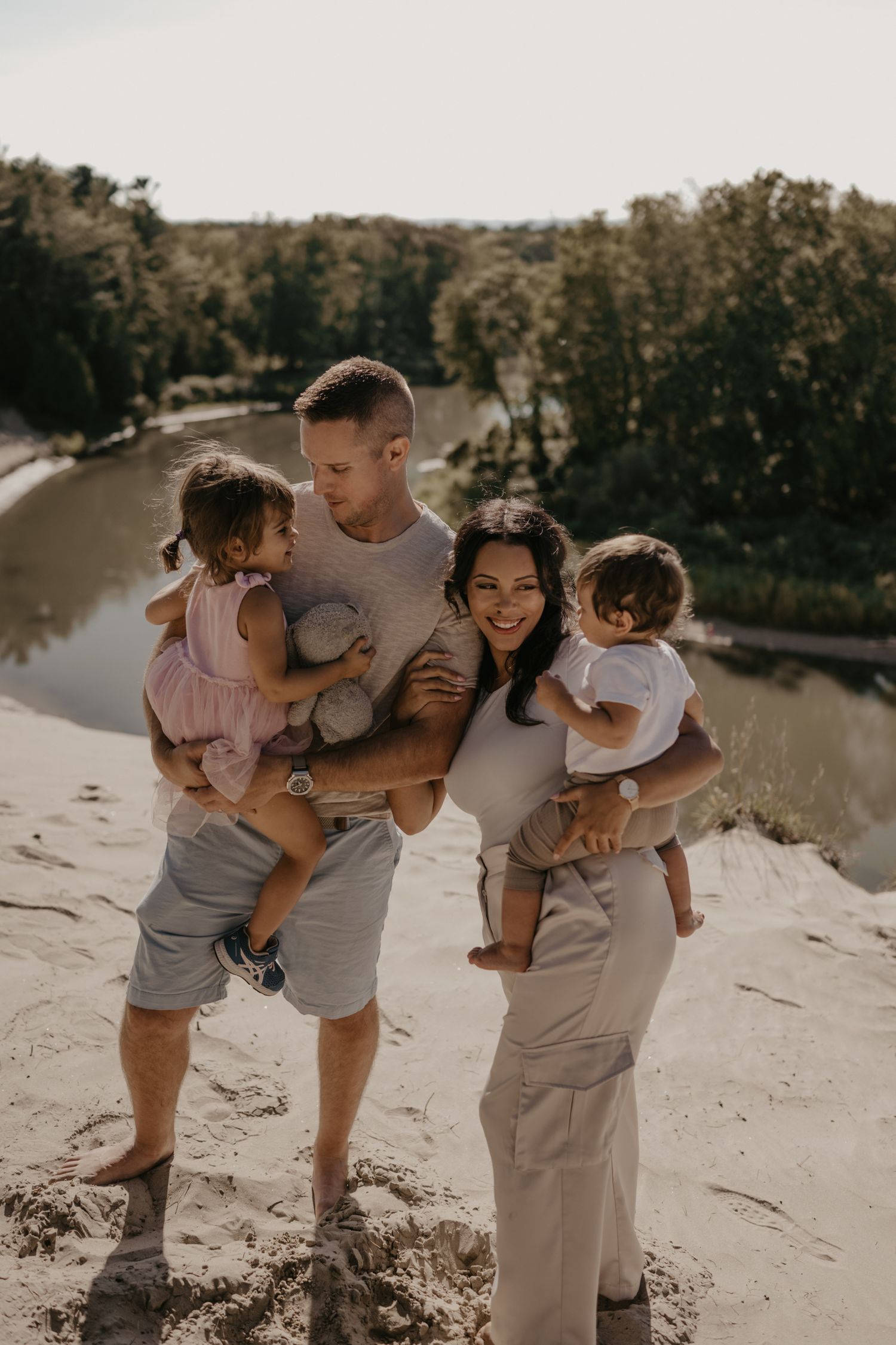 Happy family of four with two young children posing on sand dunes beside scenic river backdrop.