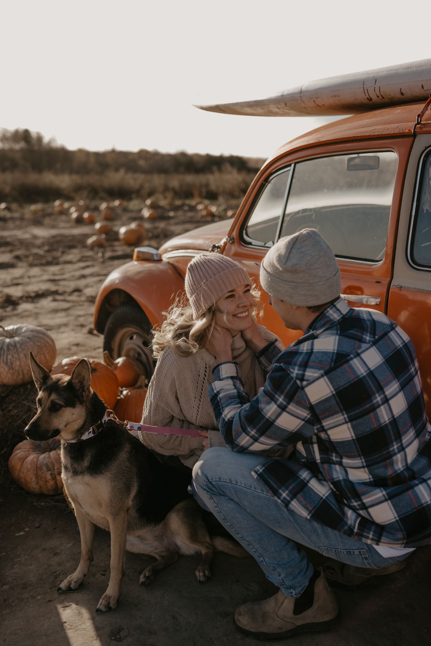Couple with German Shepherd dog sitting beside vintage orange car at pumpkin patch.