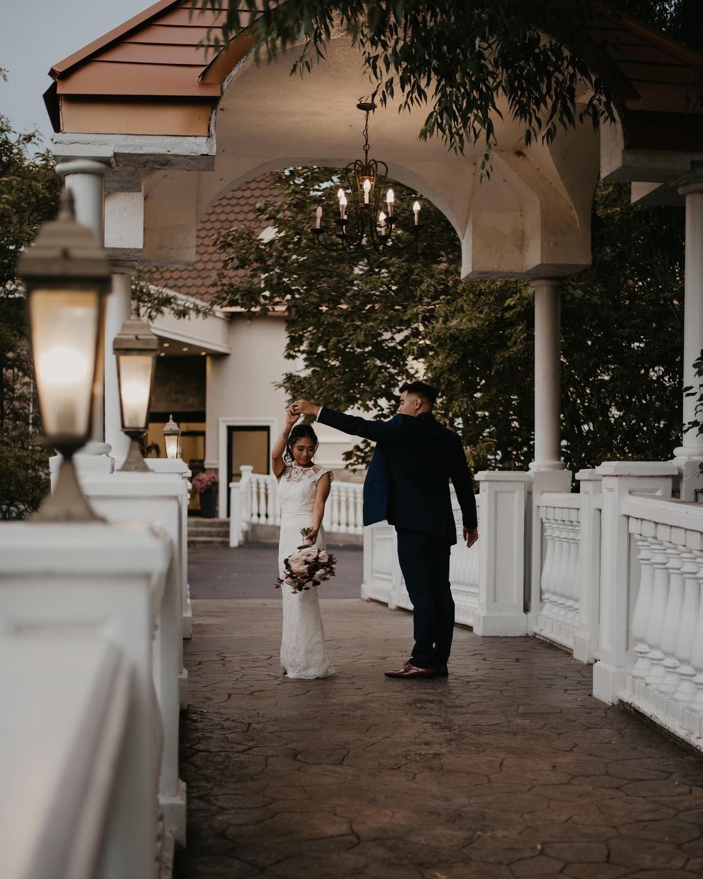 Groom spinning bride under chandelier on elegant white veranda at twilight wedding venue.