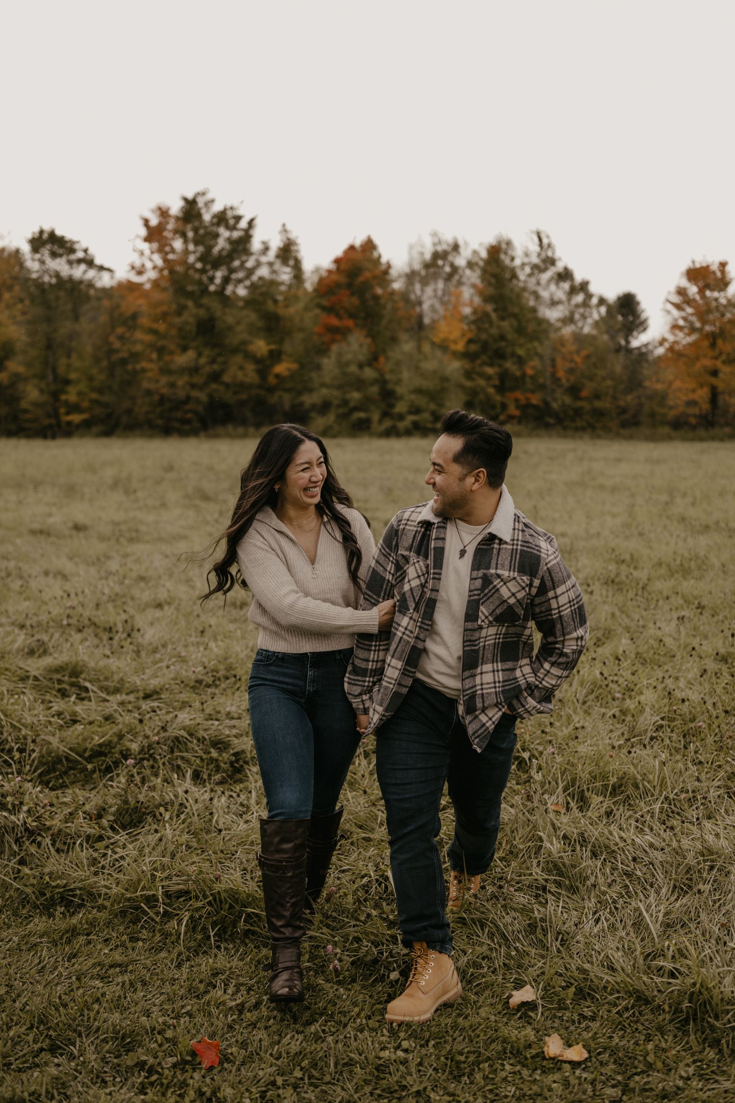 Couple laughing and walking hand in hand through a green field with colorful autumn trees in the background.