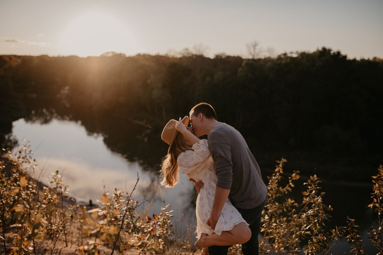 Couple sharing a romantic kiss while dipping near a sunlit river at sunset.