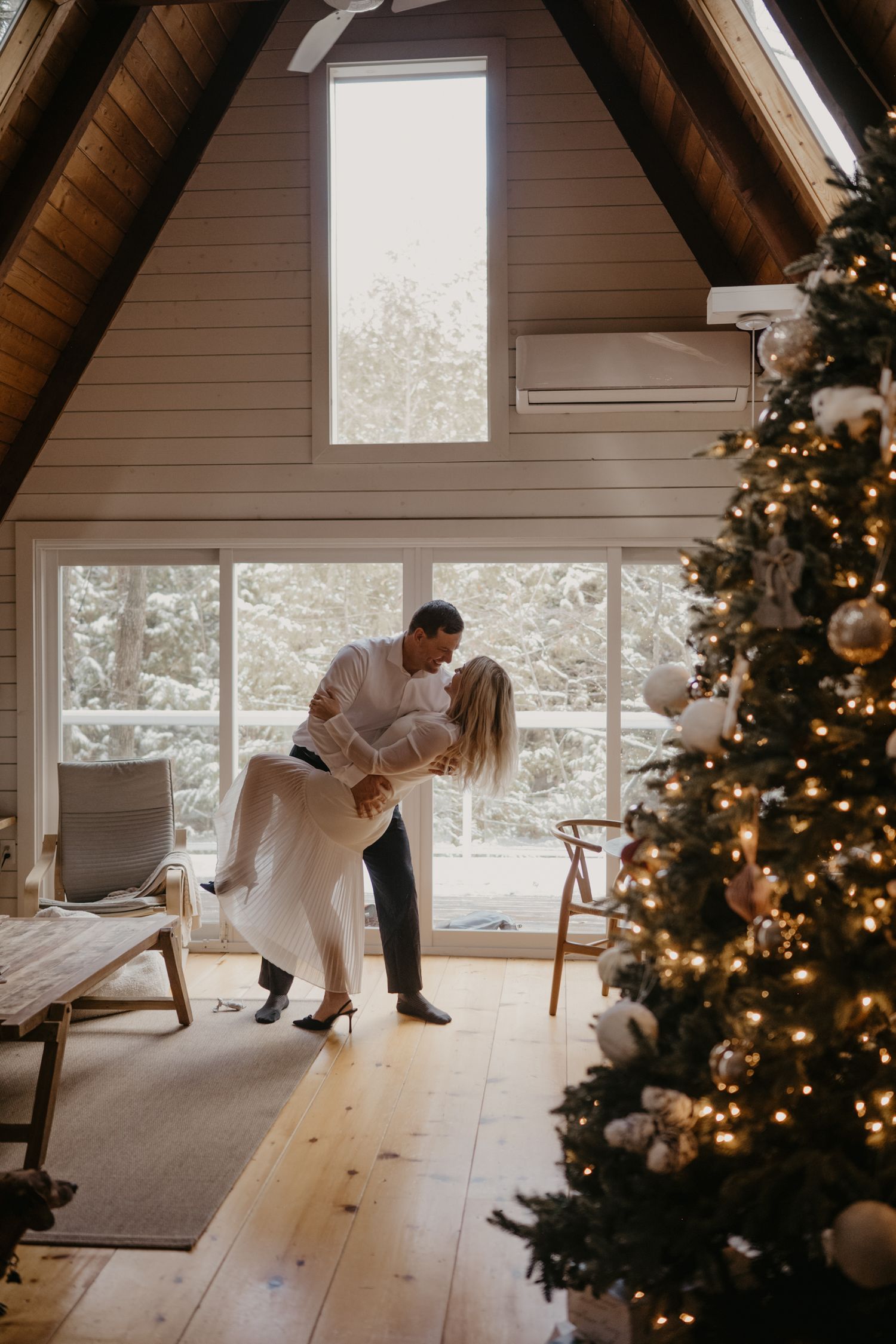 Couple sharing a romantic kiss near a decorated Christmas tree in a cozy A-frame cabin with snowy views.