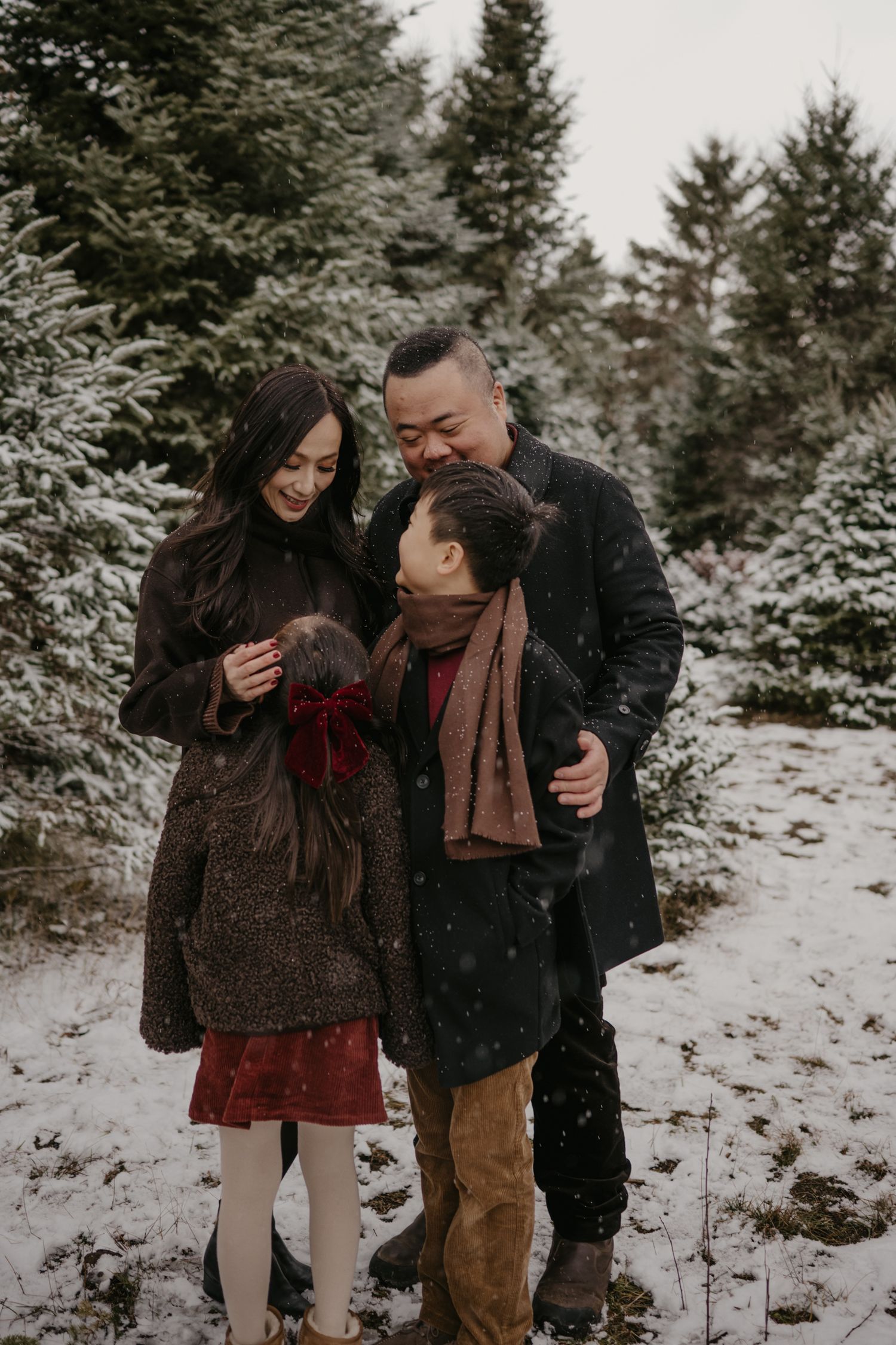 Asian family of three sharing a joyful moment in a snowy evergreen forest, dressed in festive winter attire.