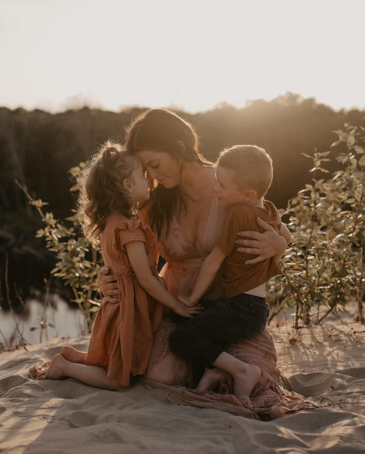 Mother hugging two young children on sandy beach during warm golden sunset light.