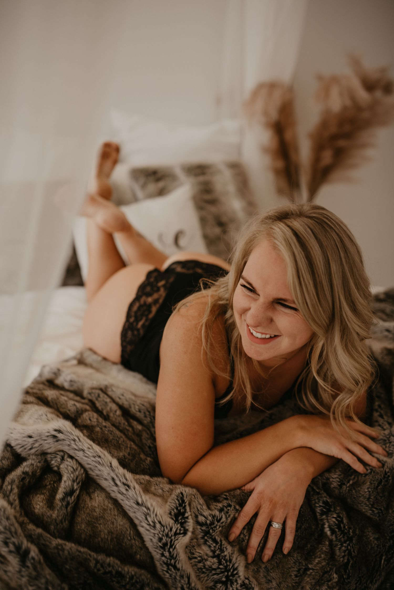 Smiling blonde woman lying on a fur blanket in black lace lingerie in a cozy bedroom.